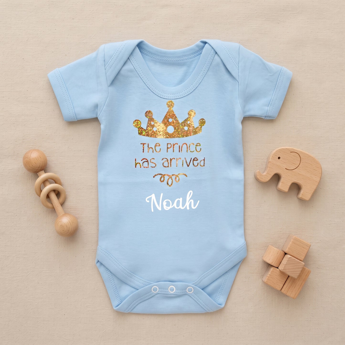 A light blue baby bodysuit laid flat on a beige fabric background, decorated with a gold glitter crown graphic and the text 'The prince has arrived' and 'Noah'. Wooden baby toys, including a rattle and blocks, are arranged around the bodysuit.
