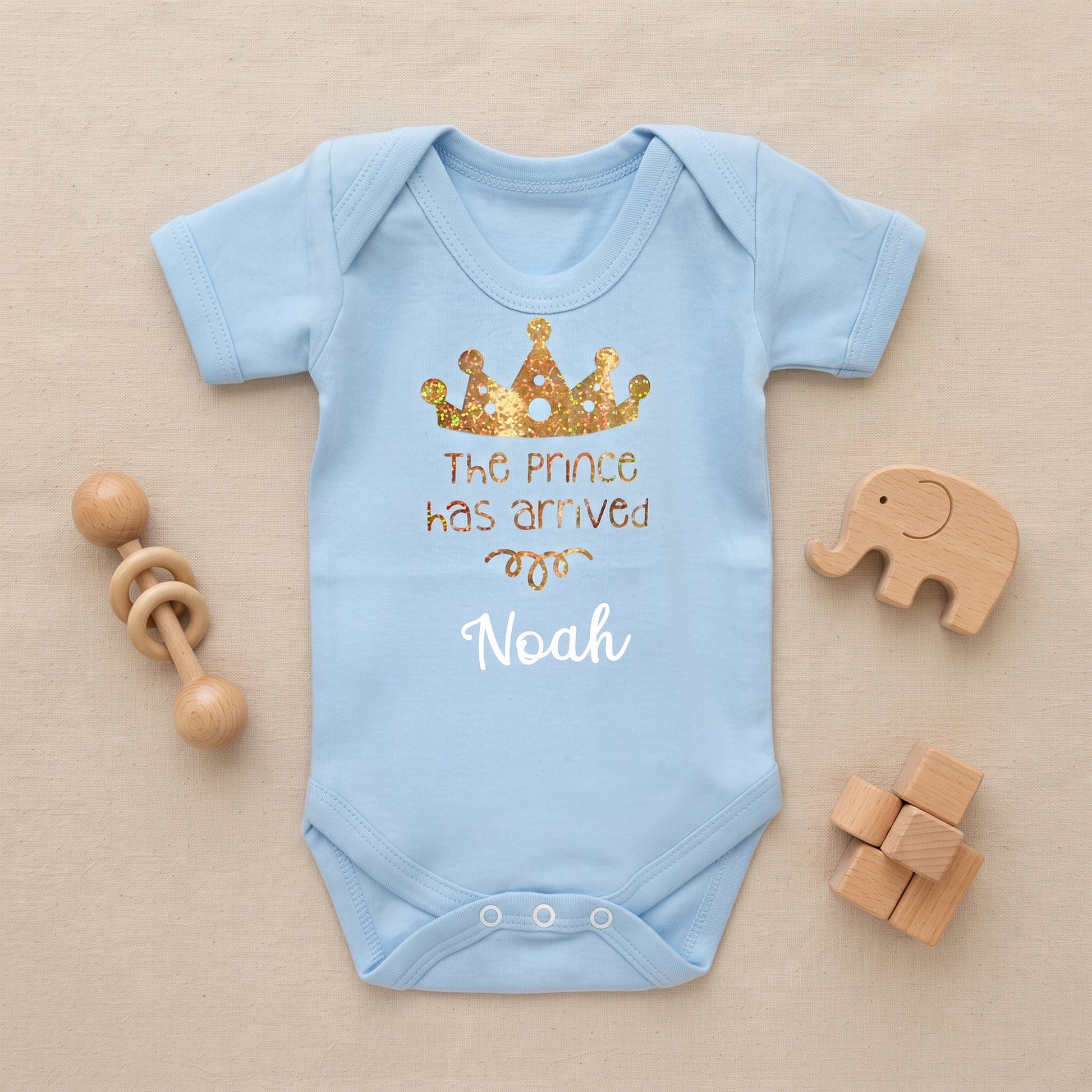 A light blue baby bodysuit laid flat on a beige fabric background, decorated with a gold glitter crown graphic and the text 'The prince has arrived' and 'Noah'. Wooden baby toys, including a rattle and blocks, are arranged around the bodysuit.