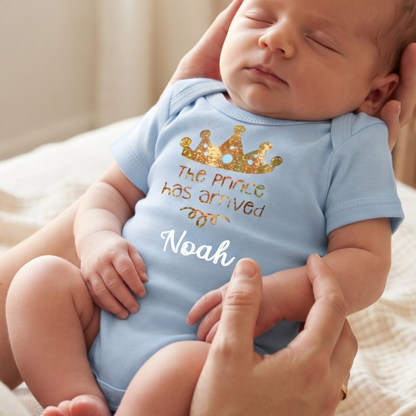 A sleeping newborn baby wearing a light blue bodysuit with a gold glitter crown and the text 'The prince has arrived Noah' in white and gold, held by adults on a soft beige blanket.