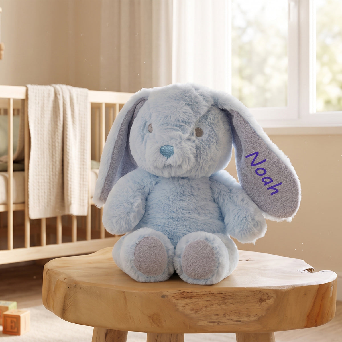 A light blue plush bunny stuffed animal with long floppy ears sits on a round wooden stool in a softly lit nursery. One ear is labeled with the name 'Noah' in purple script.