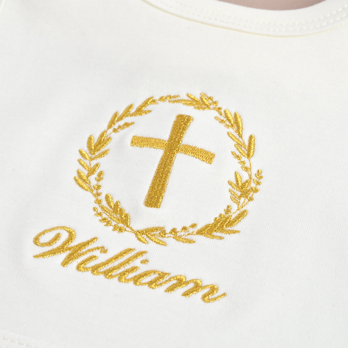 Close-up of a white garment with a gold embroidered cross inside a laurel wreath and the name 'William' in gold script below.
