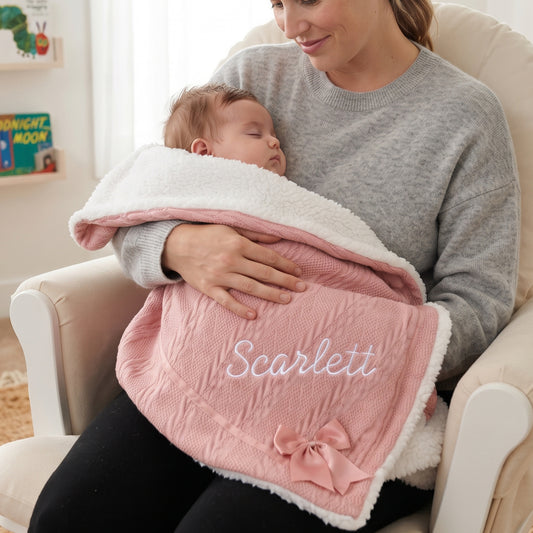 Mother holding sleeping baby wrapped in a dusty pink knitted blanket with sherpa fleece backing, personalised with the name Scarlett embroidered in white, finished with a matching bow.
