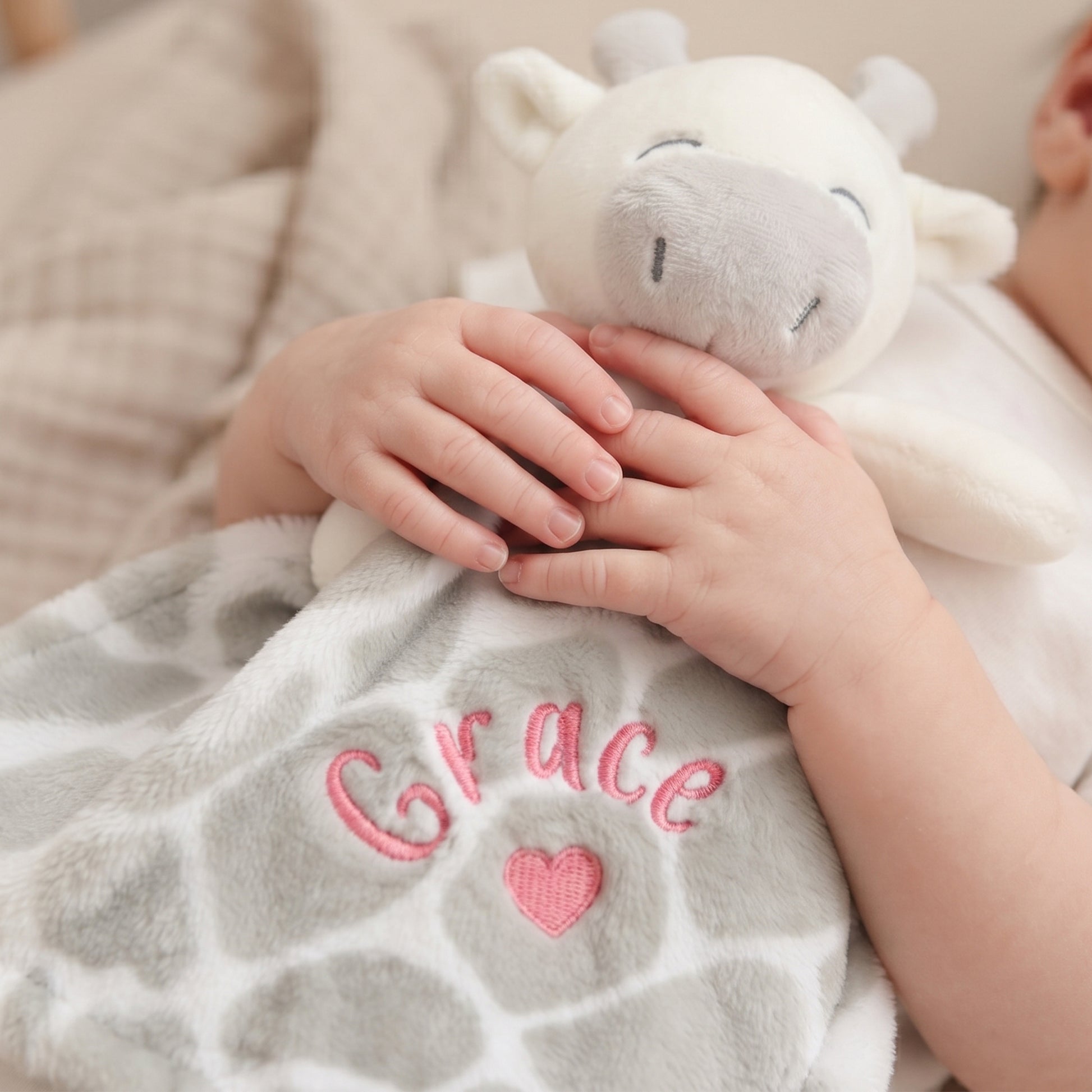 A close-up of a baby wrapped in a soft gray and white giraffe-pattern blanket with the name 'Grace' and a pink heart embroidered on it, holding a white plush cow toy.