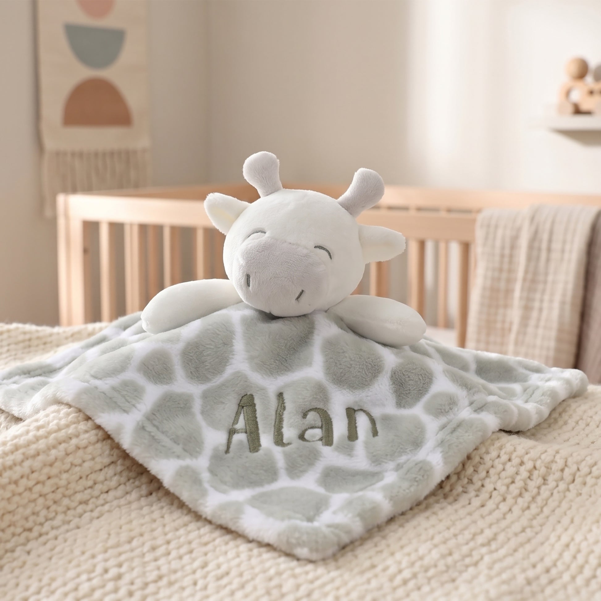 A personalized plush giraffe comforter with a soft gray and white giraffe pattern, featuring the name 'Alan' embroidered on the front, displayed on a cozy cream knit blanket in a nursery setting.