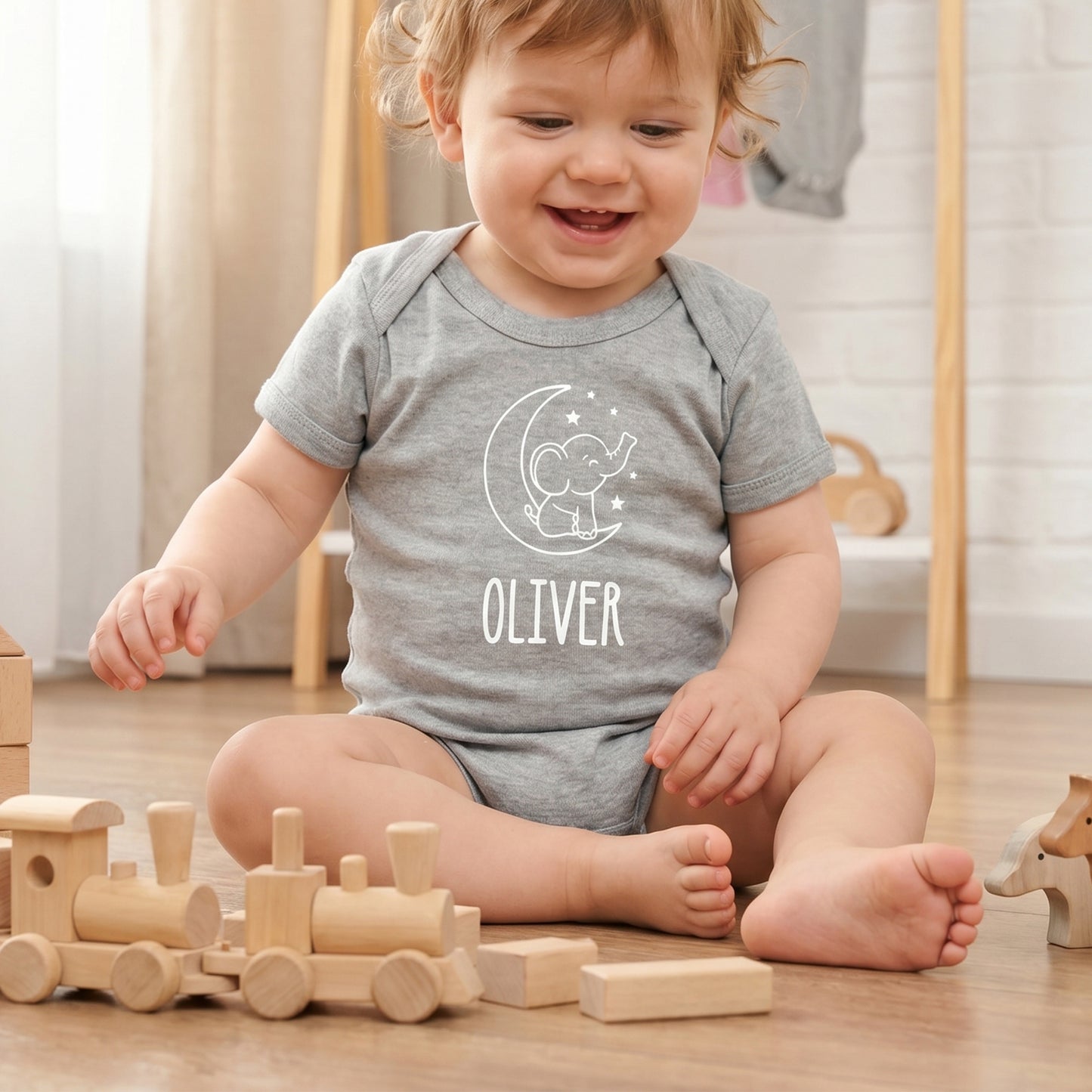 A smiling toddler sits on a wooden floor playing with wooden toy blocks, wearing a personalized heather gray short-sleeve bodysuit with a white crescent moon and elephant graphic and the name 'OLIVER' printed on the front.