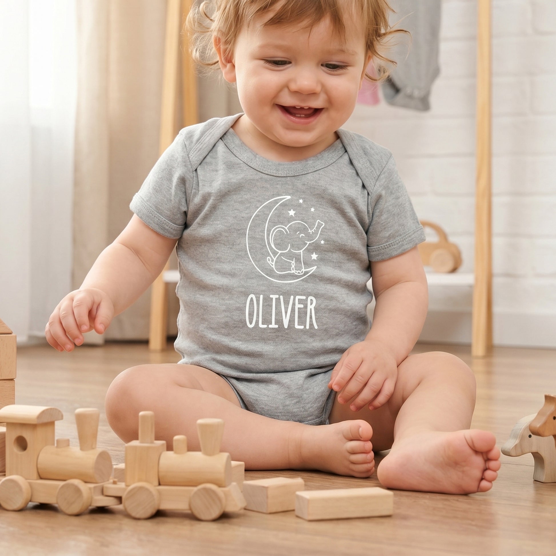 A smiling toddler sits on a wooden floor playing with wooden toy blocks, wearing a personalized heather gray short-sleeve bodysuit with a white crescent moon and elephant graphic and the name 'OLIVER' printed on the front.