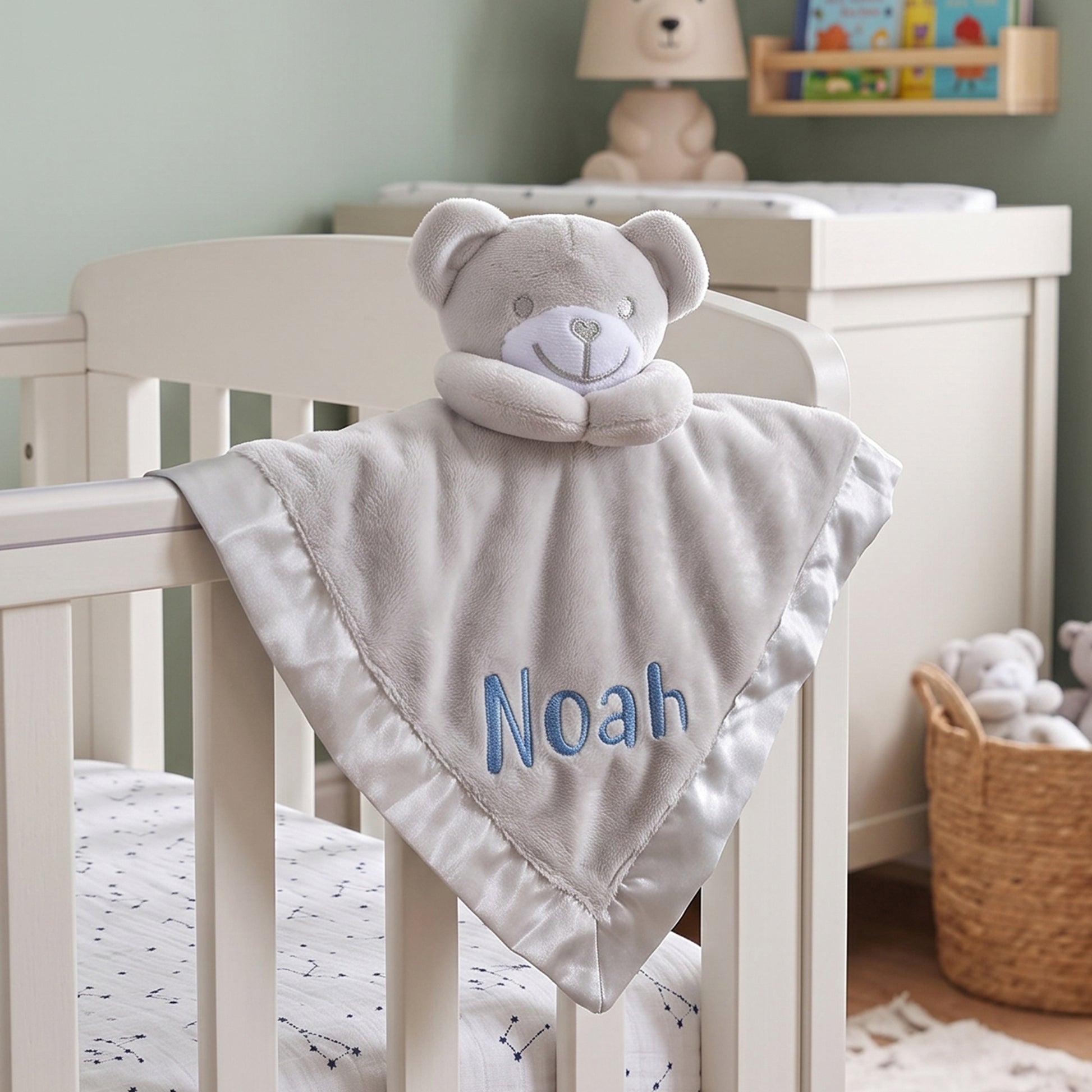 A personalized baby lovey blanket draped over a white crib, featuring a plush teddy bear head and the name 'Noah' embroidered in blue. The scene is set in a softly lit nursery with a changing table and toys in the background.