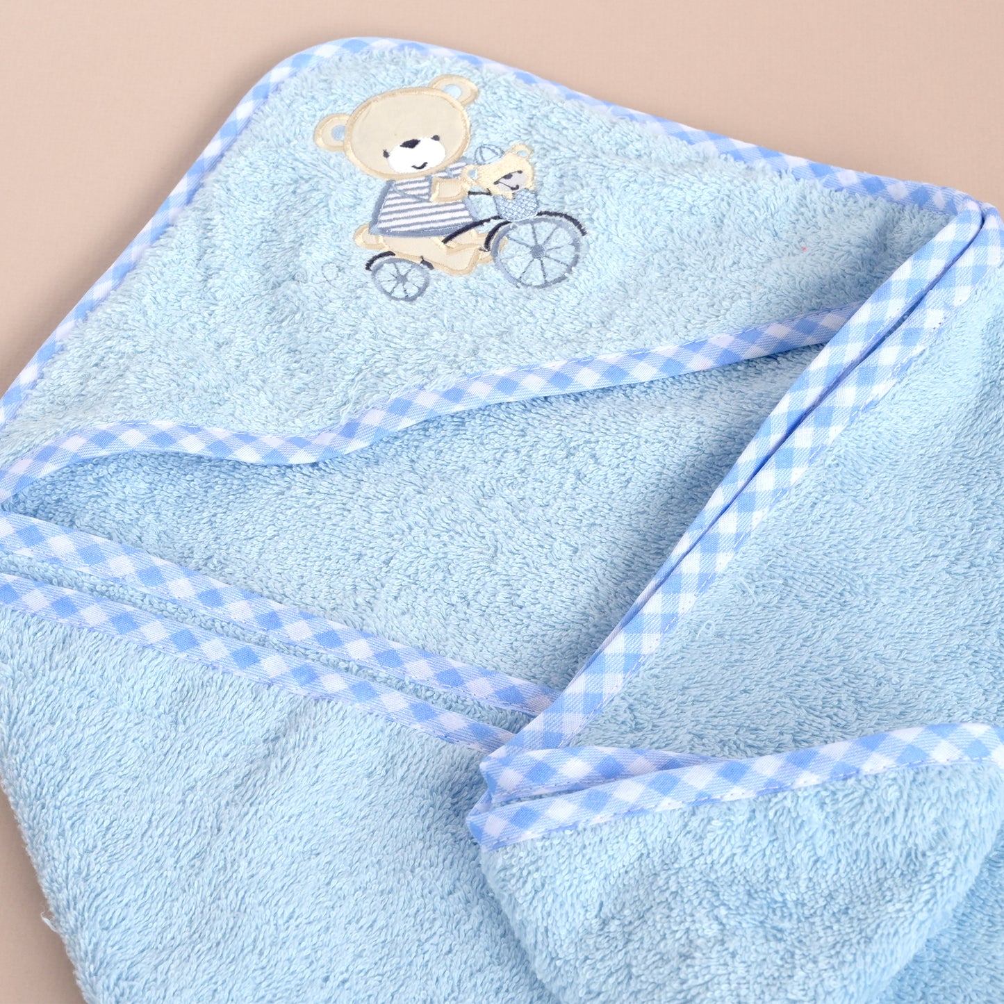 A light blue plush hooded towel with a gingham check border and an embroidered teddy bear riding a bicycle on the hood, shown on a plain beige background.