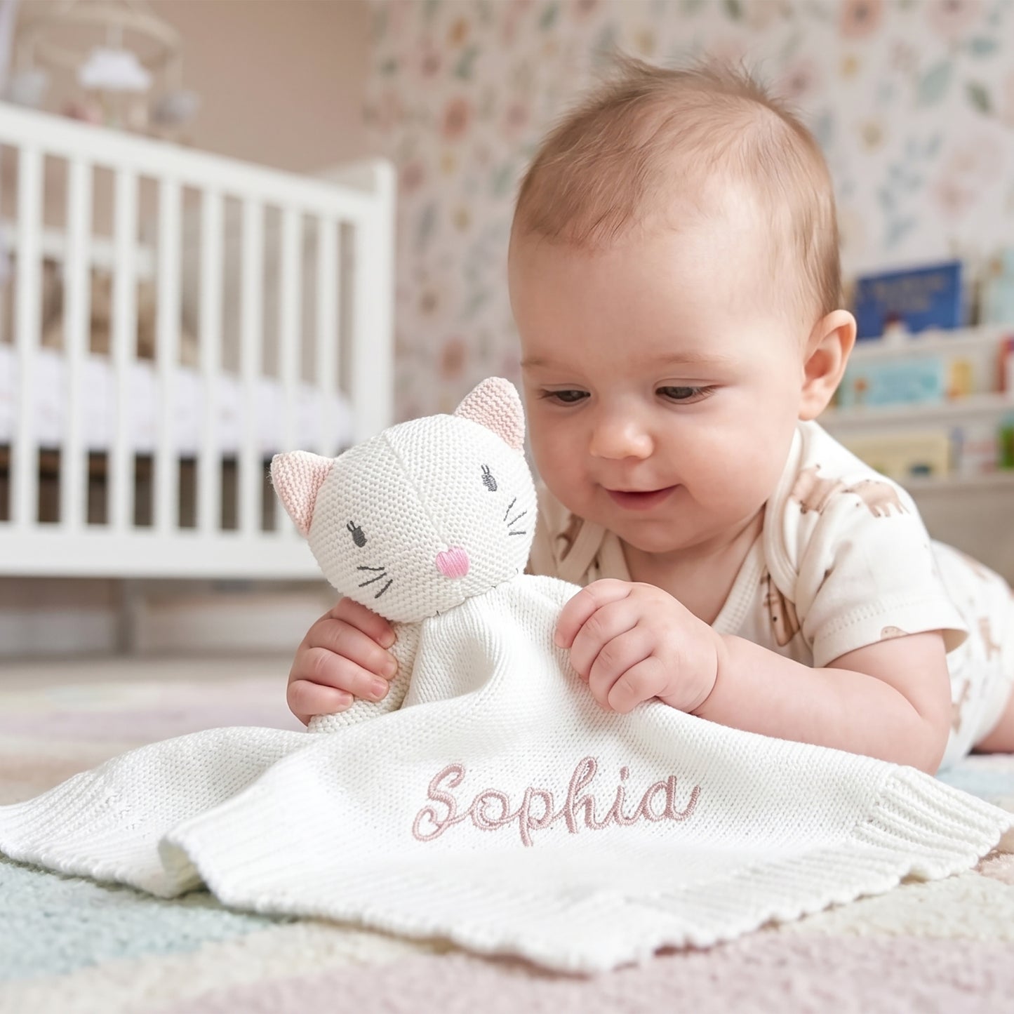 A baby lying on their stomach in a nursery, holding a white knitted cat security blanket with the name 'Sophia' embroidered in pink on the front.