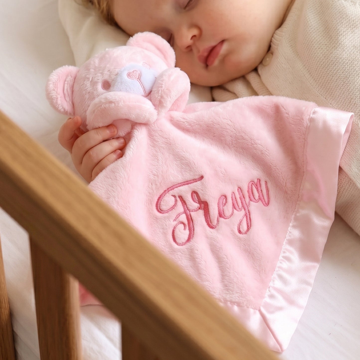 A sleeping baby in a crib holds a pink plush teddy bear comforter with the name 'Freya' embroidered in pink script on a soft pink blanket.