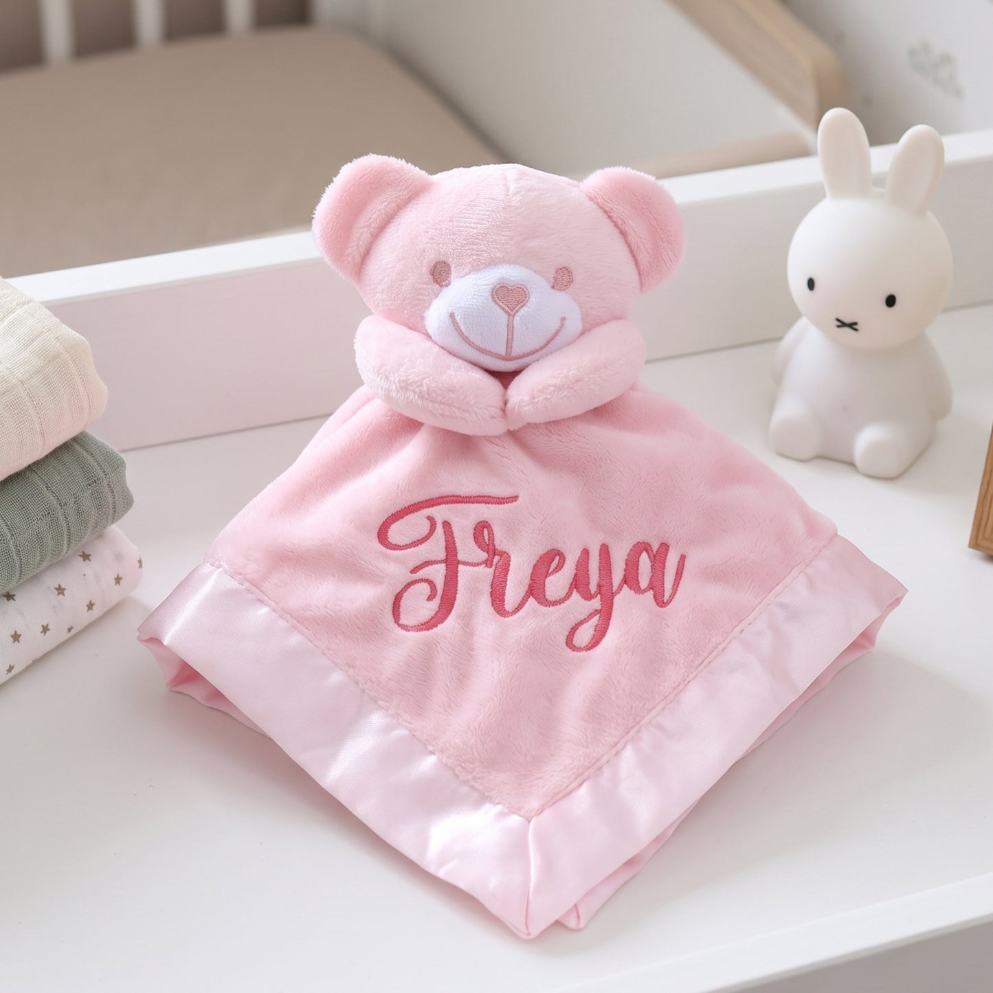 A pink plush bear security blanket with the name 'Freya' embroidered in pink script on the front, styled on a white nursery dresser next to a white rabbit figurine.