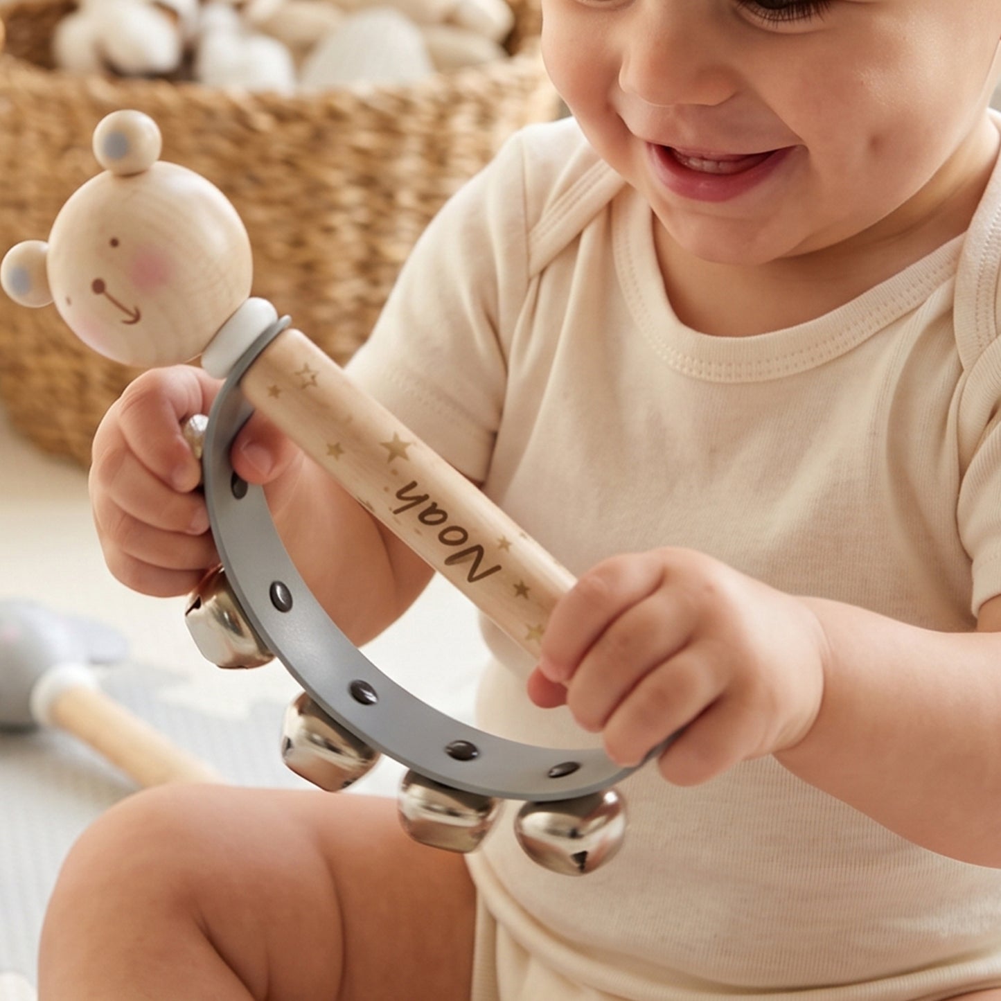 A baby in a cream onesie holds a personalized wooden toy rattle. The rattle features a bear-shaped top, a light wood handle engraved with the name 'Noah' and small gold stars, and a gray metal tambourine ring with silver bells.