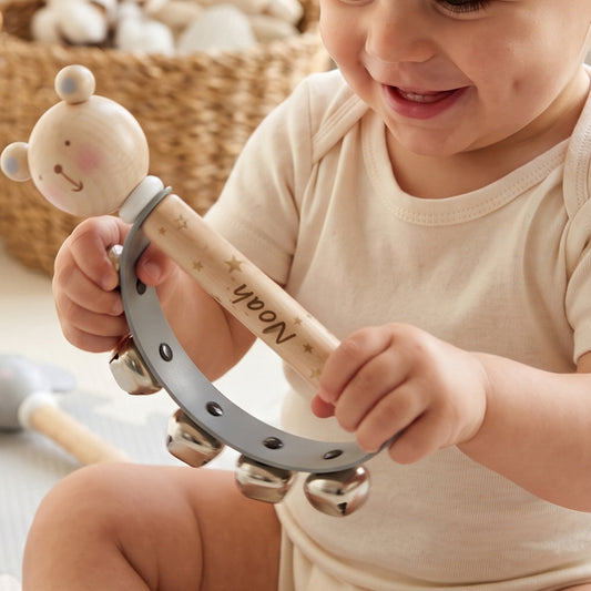 A baby in a cream onesie holds a personalized wooden toy rattle. The rattle features a bear-shaped top, a light wood handle engraved with the name 'Noah' and small gold stars, and a gray metal tambourine ring with silver bells.