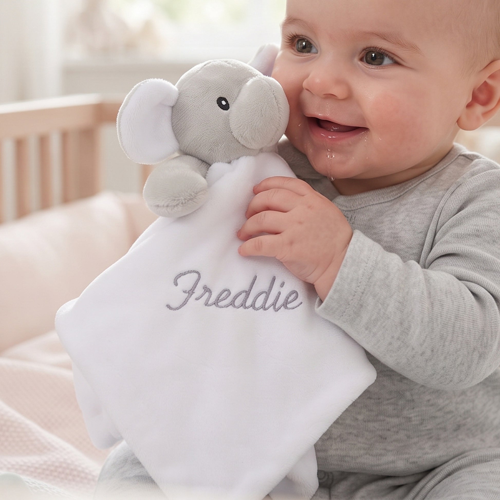 A baby in a gray long-sleeve outfit holds a white comforter with an embroidered elephant and the name 'Freddie' in gray script. The baby is smiling and looking to the side in a softly lit nursery setting.
