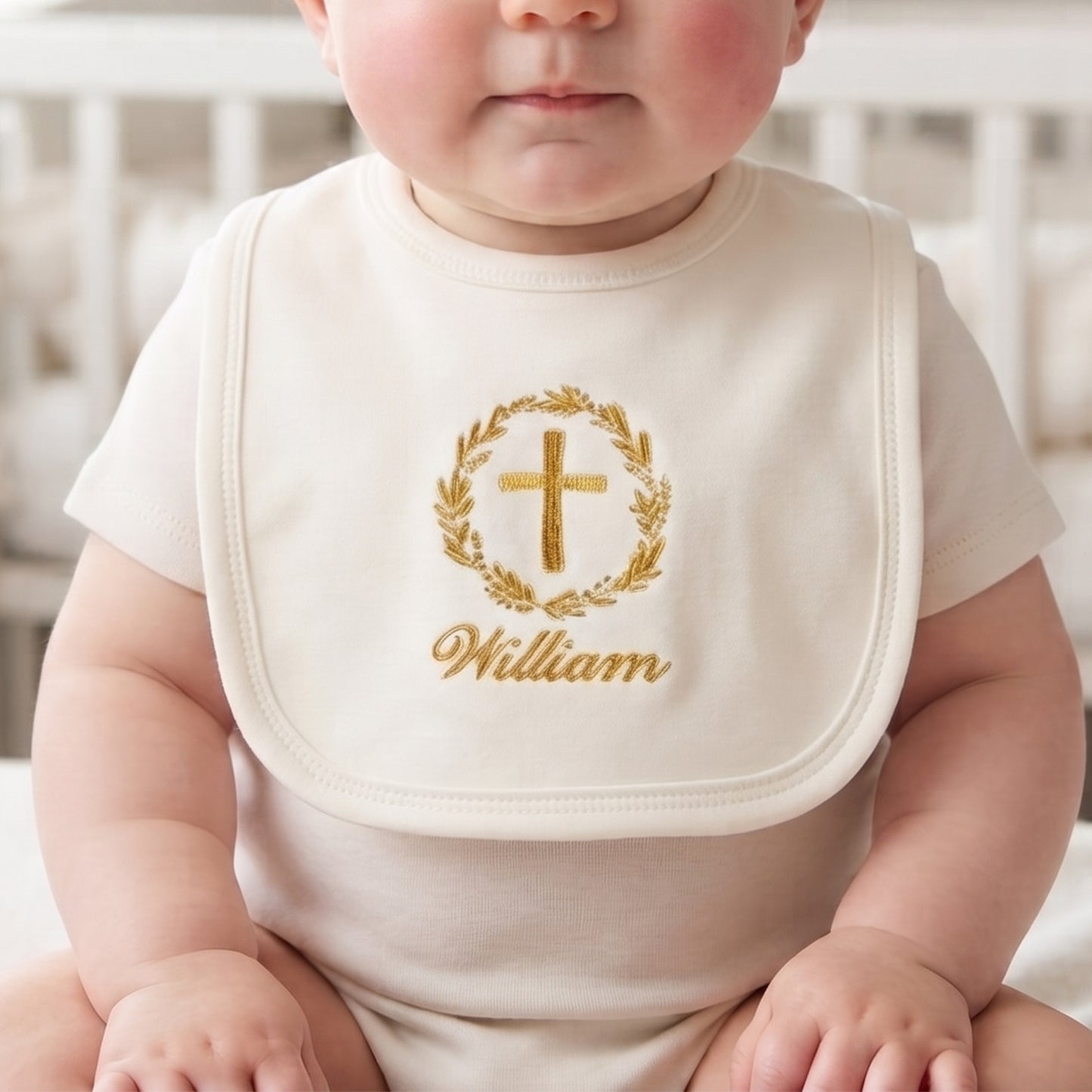 Cream cotton christening bib with gold embroidered wreath and cross, personalised with baby's name, on a wooden surface