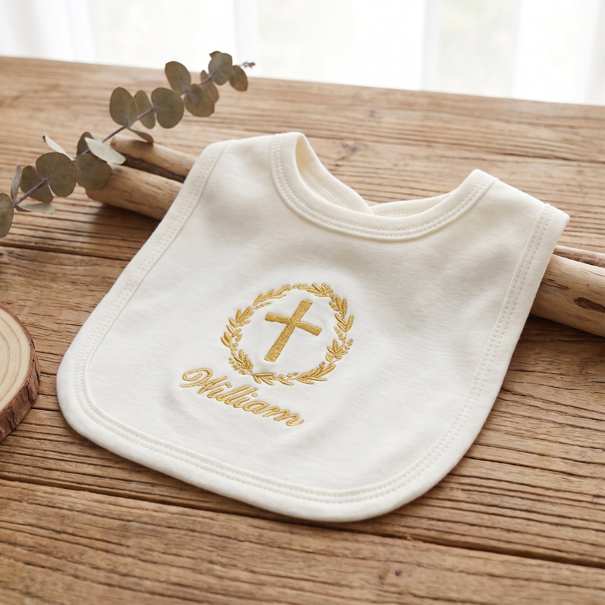 An off-white personalized baby bib with gold embroidery featuring a cross, a laurel wreath, and the name 'William', displayed on a rustic wooden surface with eucalyptus leaves.