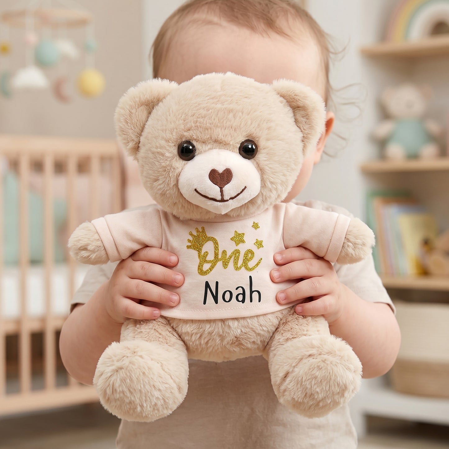 A baby holding a plush teddy bear in a nursery setting. The teddy bear is cream-colored with a light pink shirt printed with 'One Noah' in gold and black text.