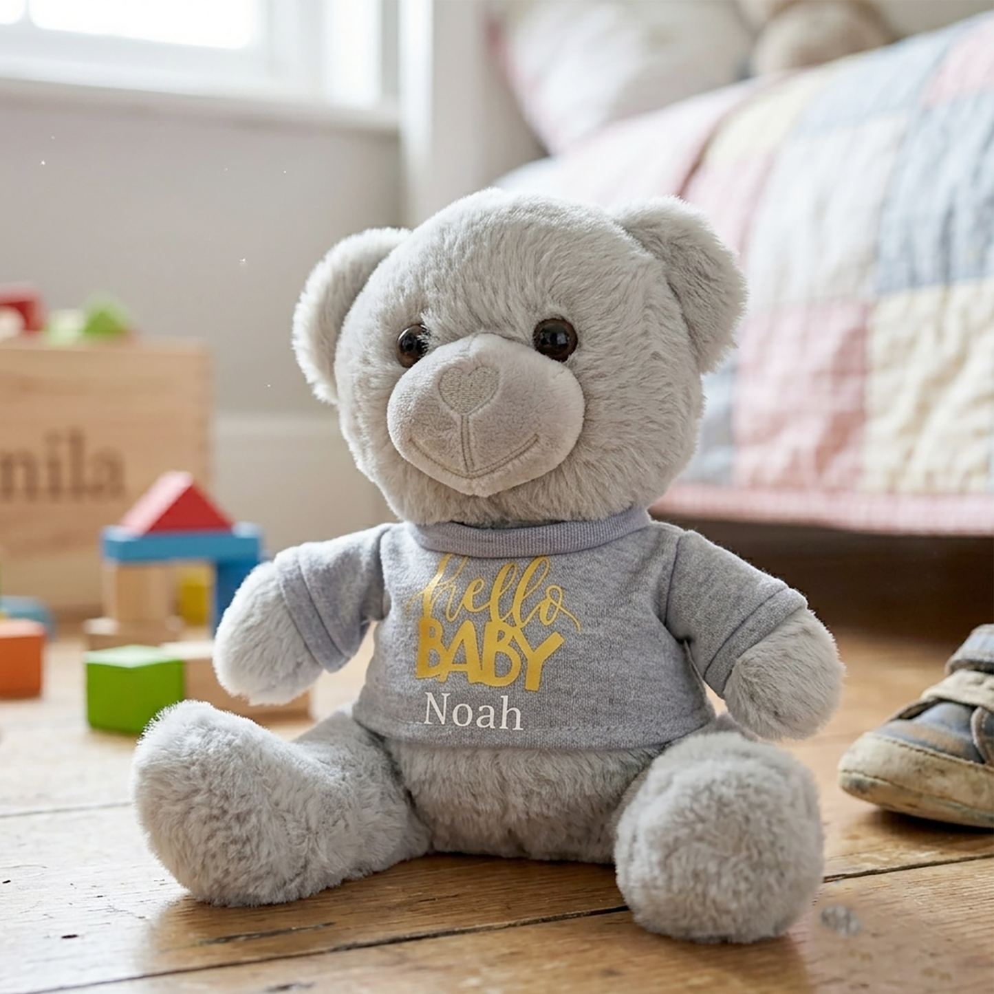Grey teddy bear soft toy wearing a grey t-shirt printed with "Hello Baby" in gold, personalised with the name Noah, sitting on a cot mattress next to wooden alphabet blocks.
