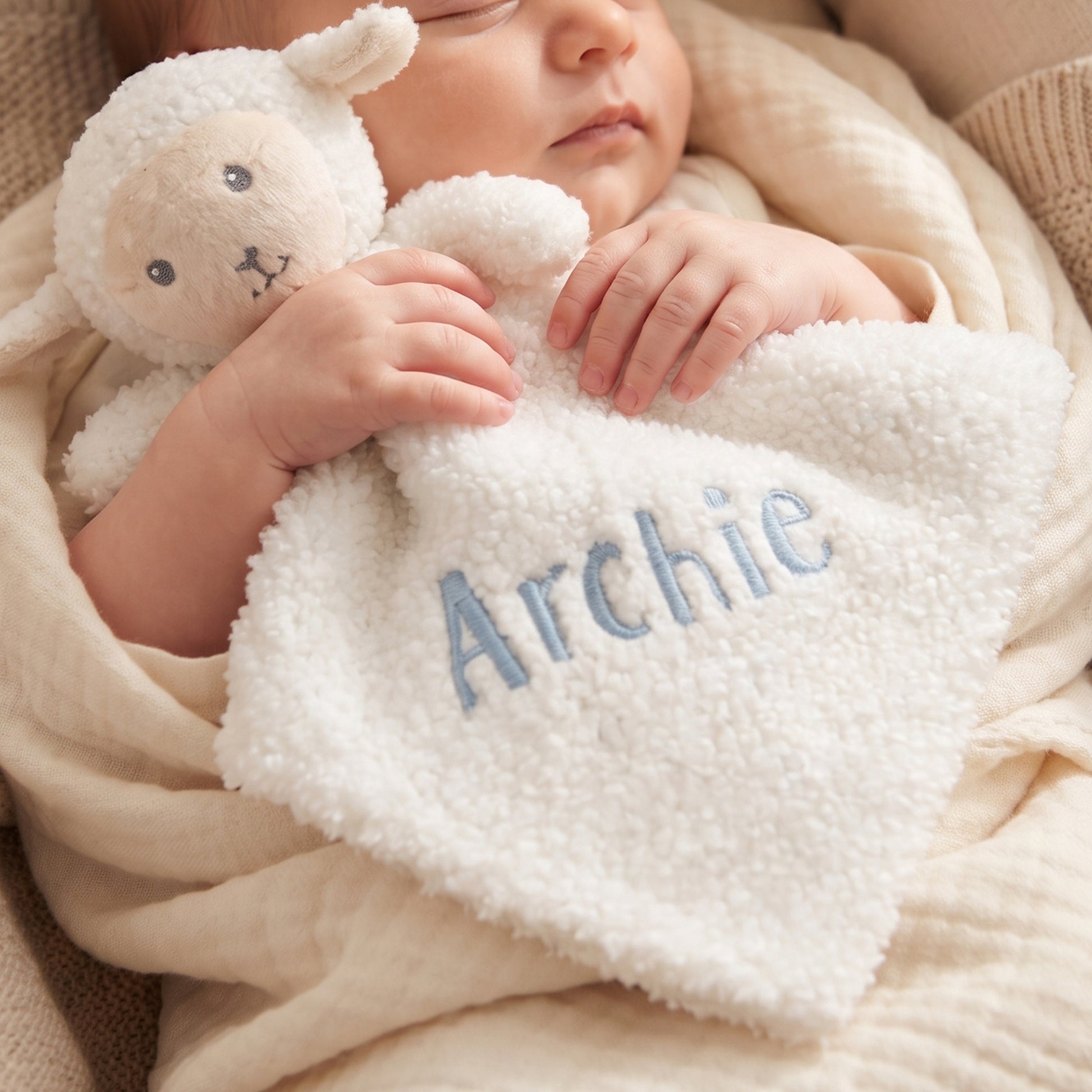 A sleeping baby wrapped in a soft cream blanket, holding a plush sheep lovey with the name 'Archie' embroidered in light blue on the front.