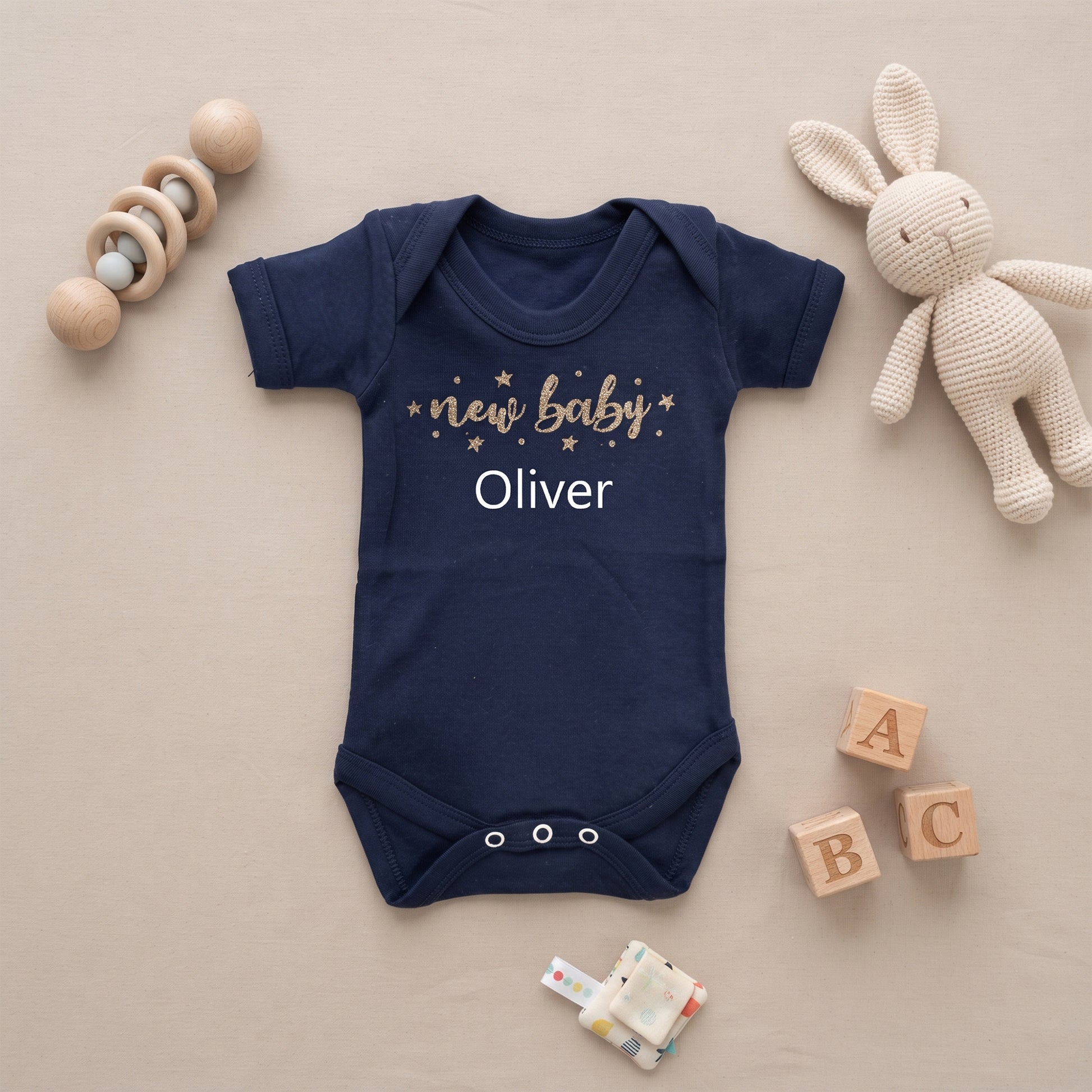 A navy blue short-sleeve baby bodysuit with 'new baby Oliver' printed on the chest in white and gold, styled in a flat lay with wooden toys, a knitted bunny doll, and alphabet blocks on a beige background.
