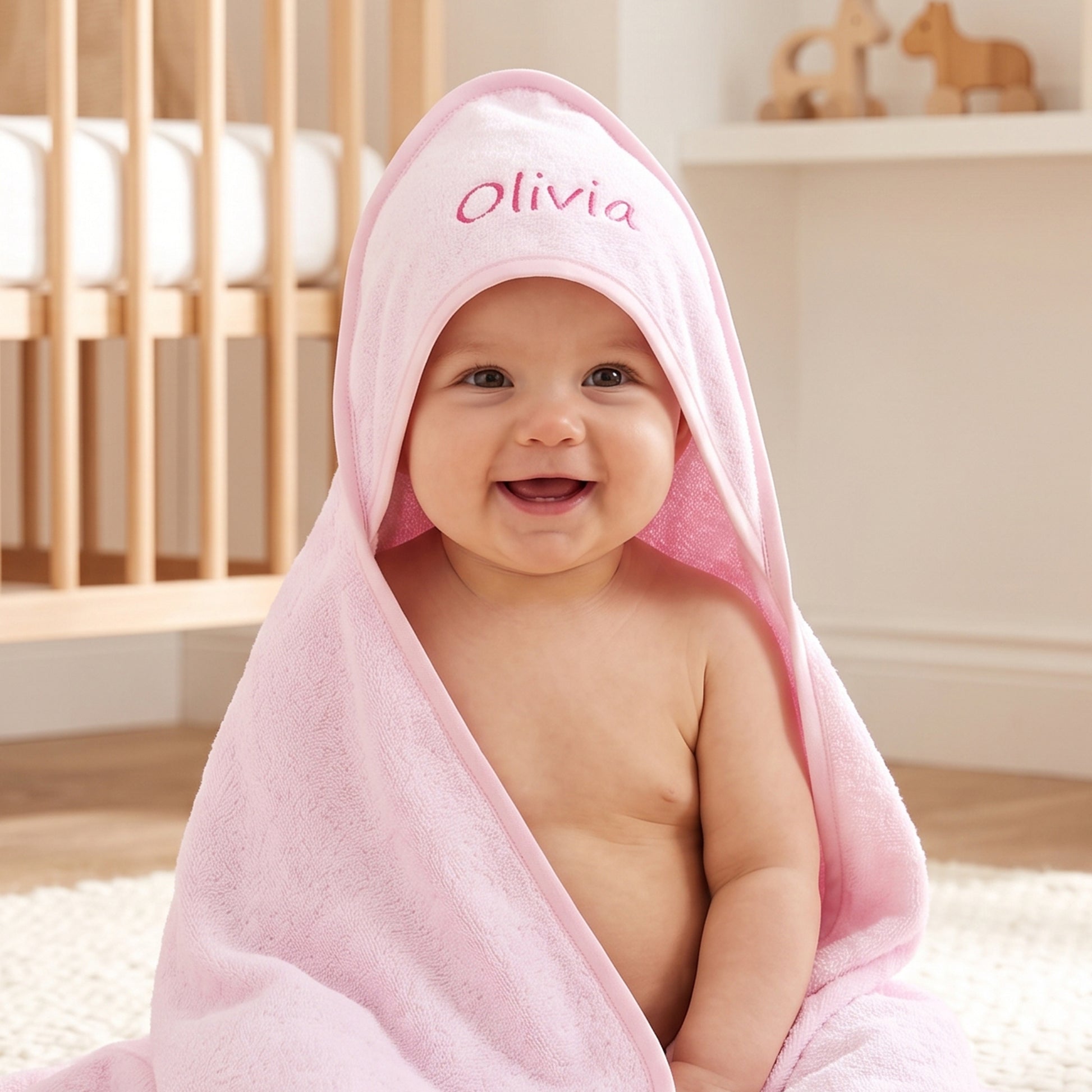 Bright pink hooded baby towel in cotton terry, personalised with the name Olivia embroidered in a deeper pink, worn by a smiling baby sitting on a rug.