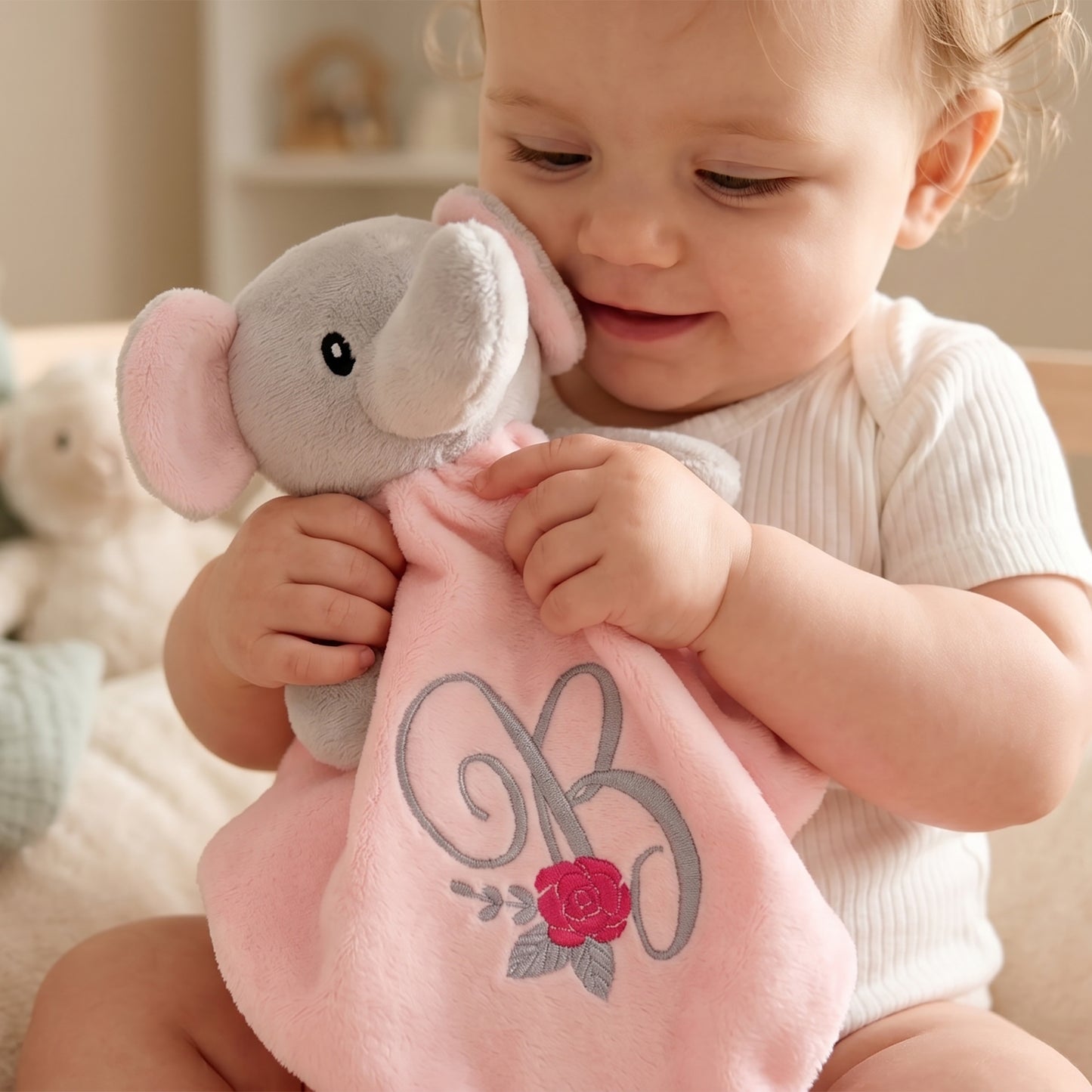 A baby wearing a white ribbed bodysuit holds and cuddles a plush elephant comforter. The comforter is light pink with a gray elephant head and features an embroidered gray monogram 'R' and a red rose on the front.