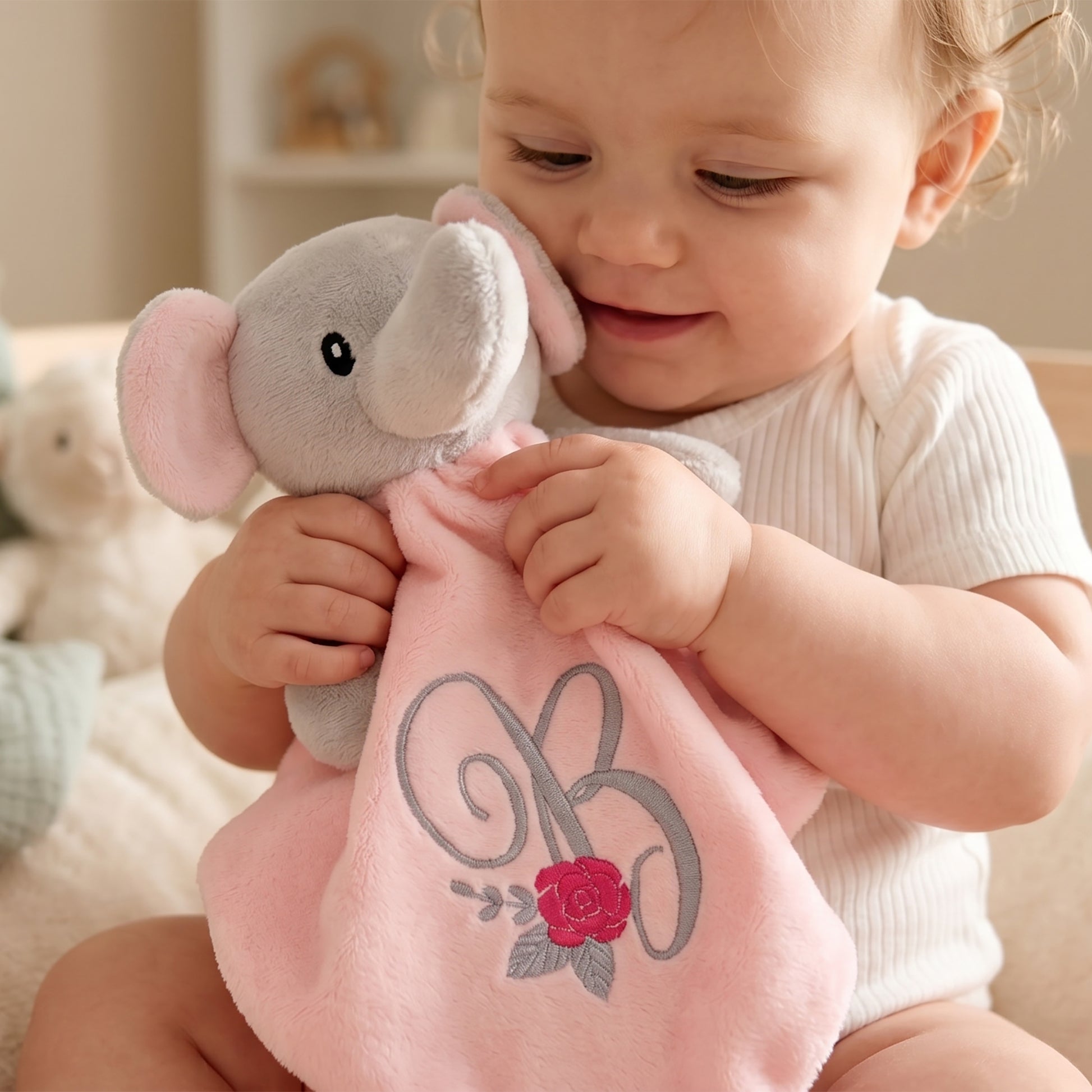 A baby wearing a white ribbed bodysuit holds and cuddles a plush elephant comforter. The comforter is light pink with a gray elephant head and features an embroidered gray monogram 'R' and a red rose on the front.