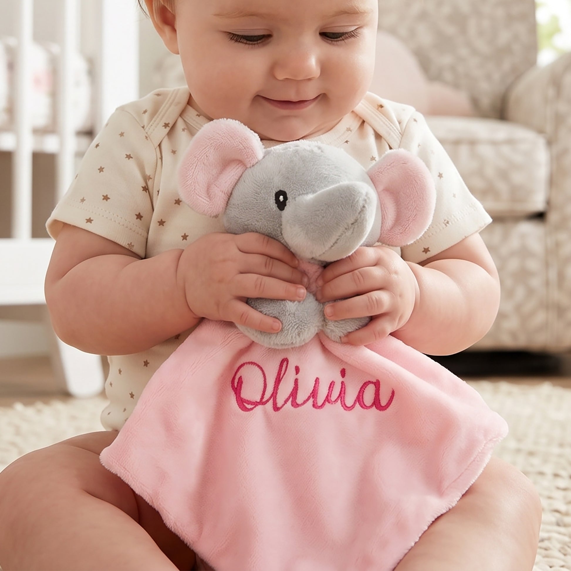 A baby sitting on a carpeted floor holds a personalized soft toy comforter. The plush toy features a gray elephant head with pink ears and a pink blanket body embroidered with the name 'Olivia' in pink script.