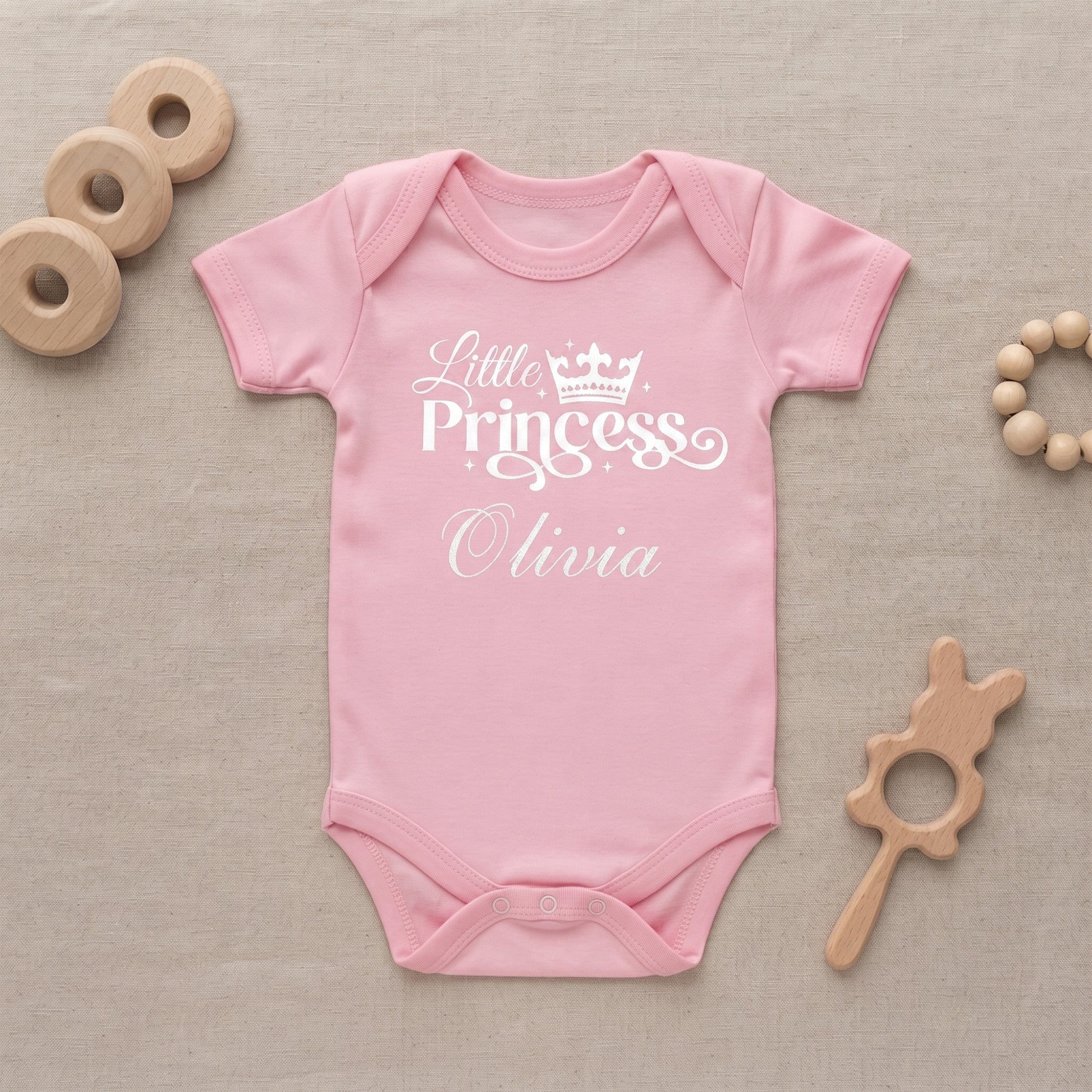 A pink short-sleeve baby bodysuit with 'Little Princess Olivia' printed in white on the front, laid flat on a beige fabric background with wooden baby toys around it.