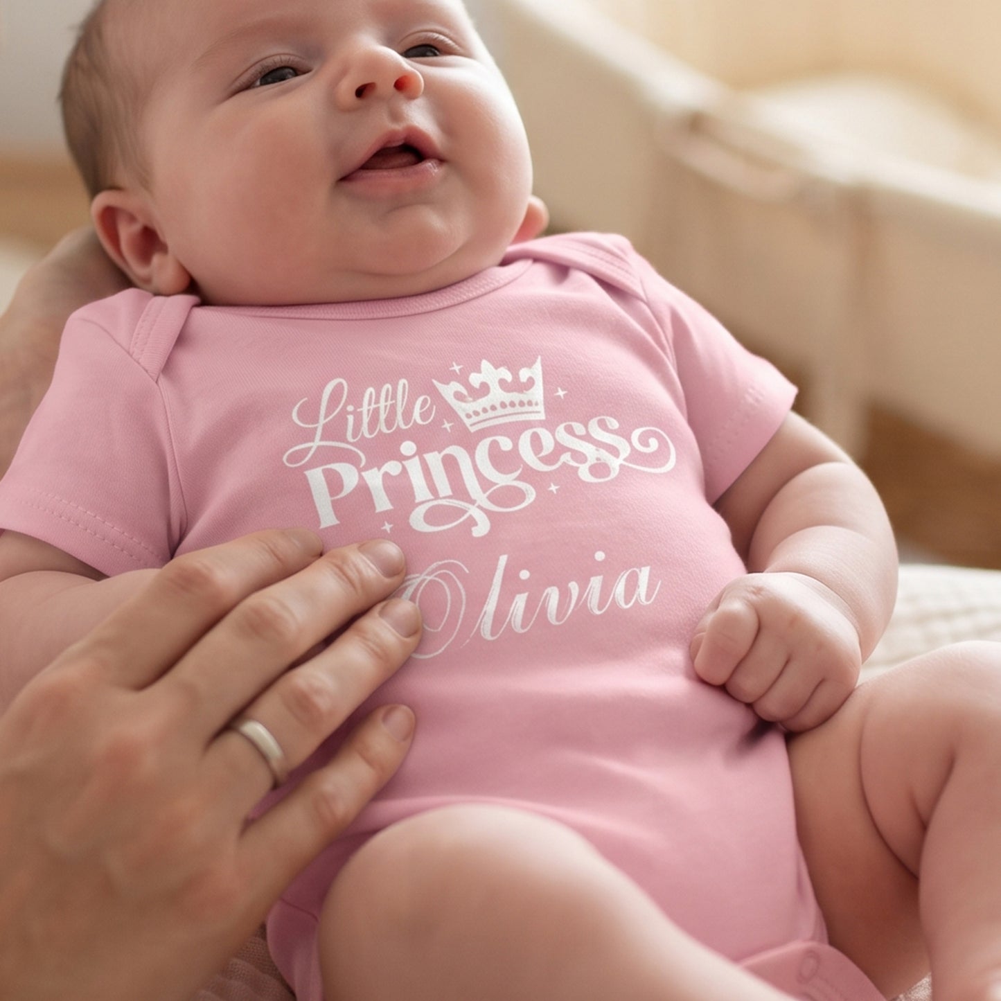 A close-up lifestyle shot of a baby wearing a light pink short-sleeve bodysuit with 'Little Princess Olivia' printed in white script and a crown graphic. An adult hand is gently holding the baby, and the background is a soft, warm, neutral indoor setting.