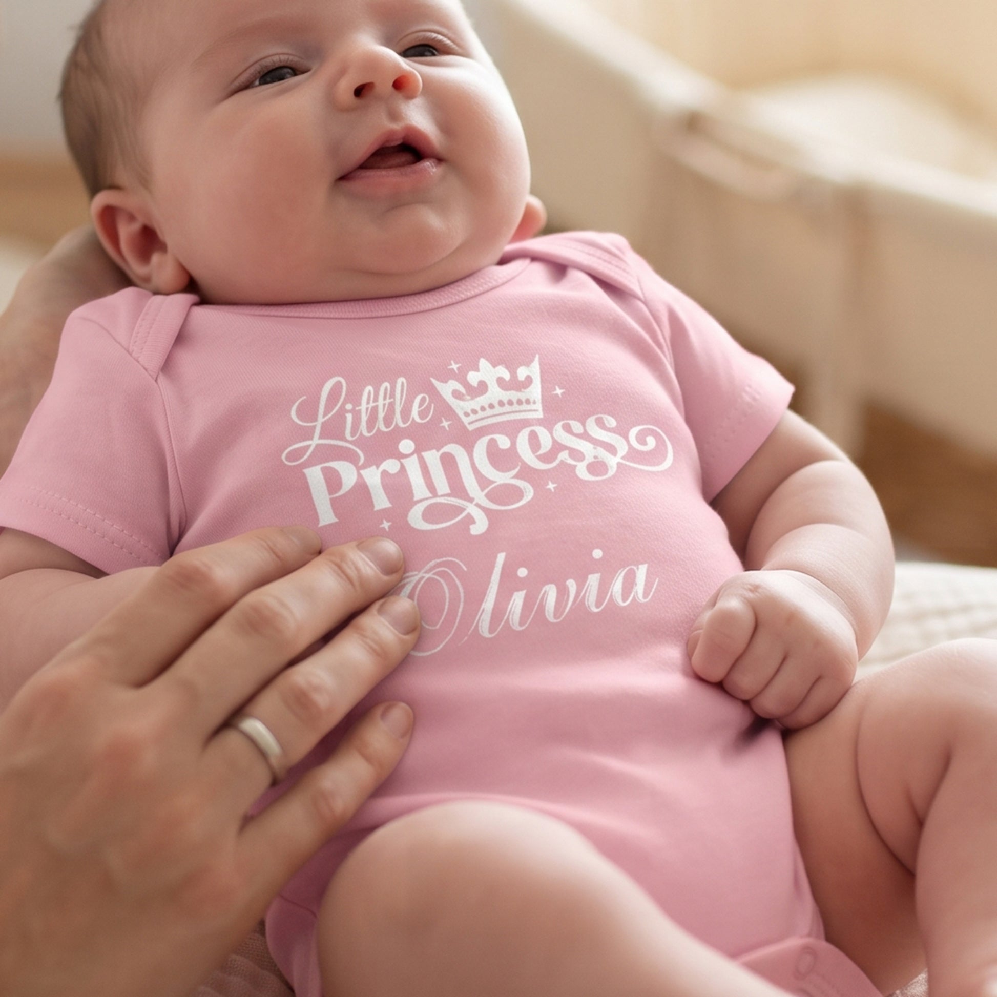 A close-up lifestyle shot of a baby wearing a light pink short-sleeve bodysuit with 'Little Princess Olivia' printed in white script and a crown graphic. An adult hand is gently holding the baby, and the background is a soft, warm, neutral indoor setting.