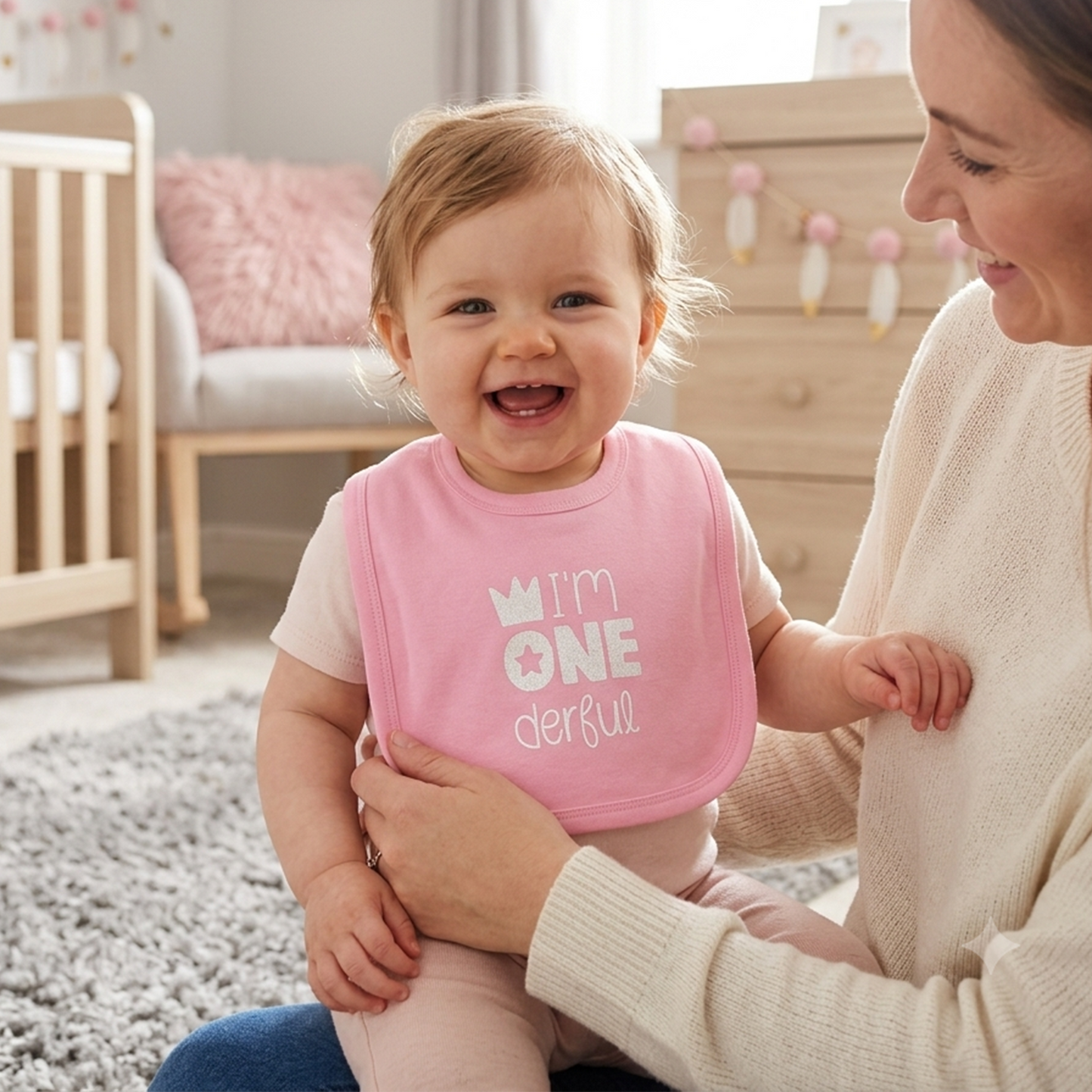 A baby wearing a pink bib with the text 'I'm ONE d*erful' sits on a mother's lap in a softly lit nursery. The bib has a white crown and star graphic.