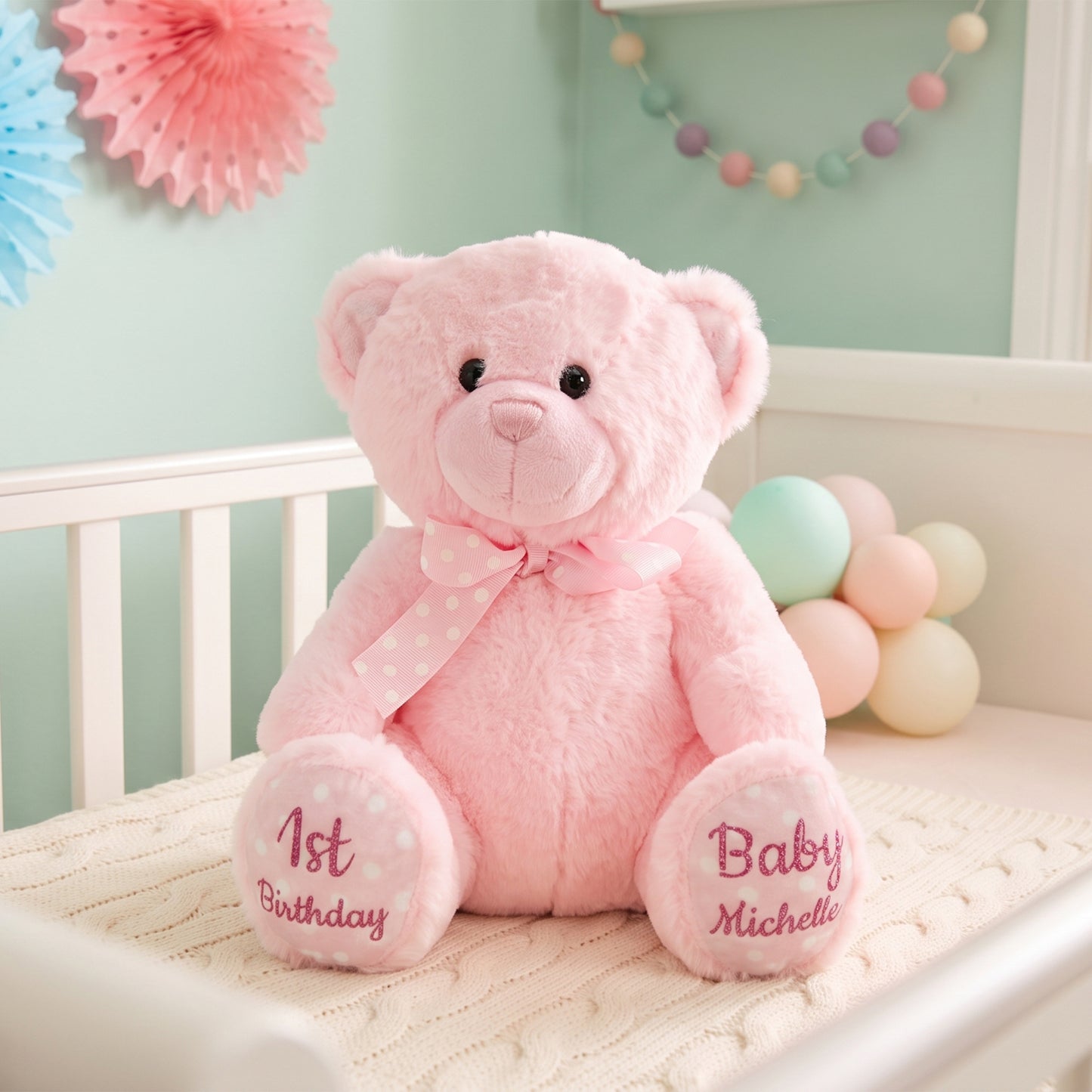 A pink plush teddy bear sits on a white crib in a pastel nursery. The bear has embroidered pink text on its feet reading '1st Birthday' and 'Baby Michelle,' plus a pink polka-dot bow around its neck.