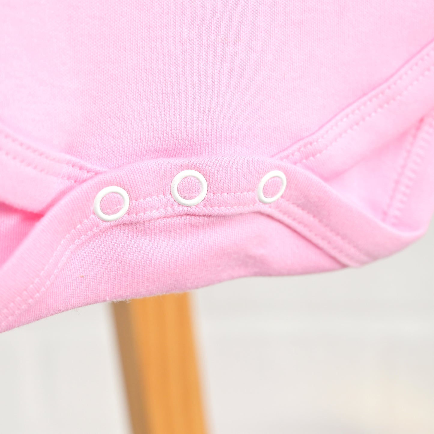 Close-up of a pink baby bodysuit or romper with three white snap closures along the neckline, shown against a soft light background with a blurred wooden stand visible below.