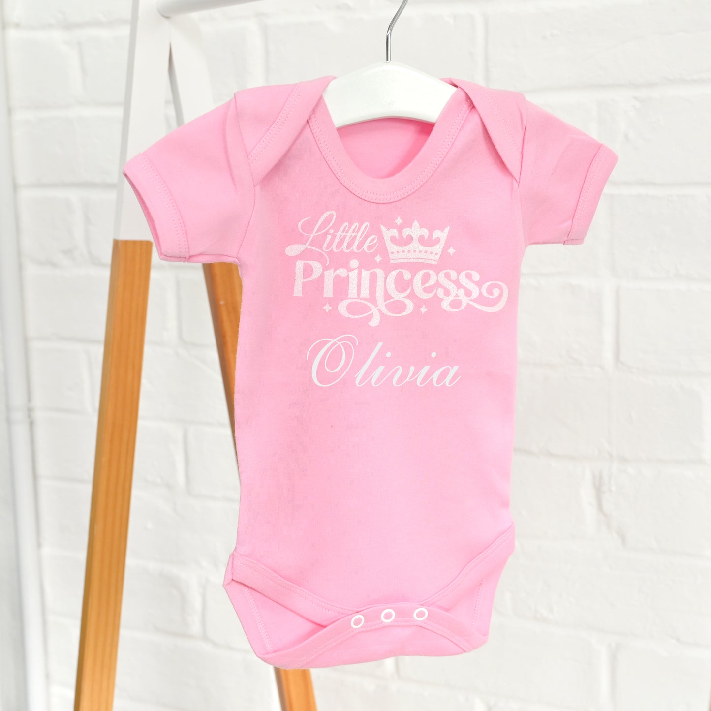 A pink short-sleeve baby bodysuit hanging on a white hanger against a white brick wall, with white printed text reading 'Little Princess Olivia' and a small crown graphic on the chest.