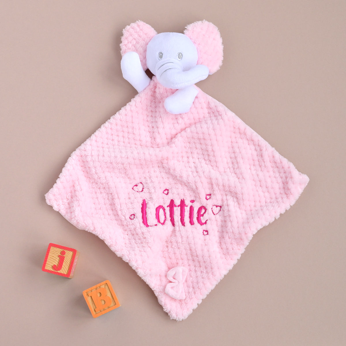 Pink honeycomb-textured comforter with a white elephant head, personalised with the name "Lottie" and small pink hearts in embroidery, displayed on a stack of folded muslins.