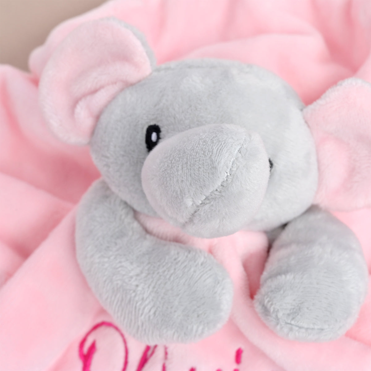 A close-up of a soft plush elephant toy resting on a pink blanket with embroidered lettering.