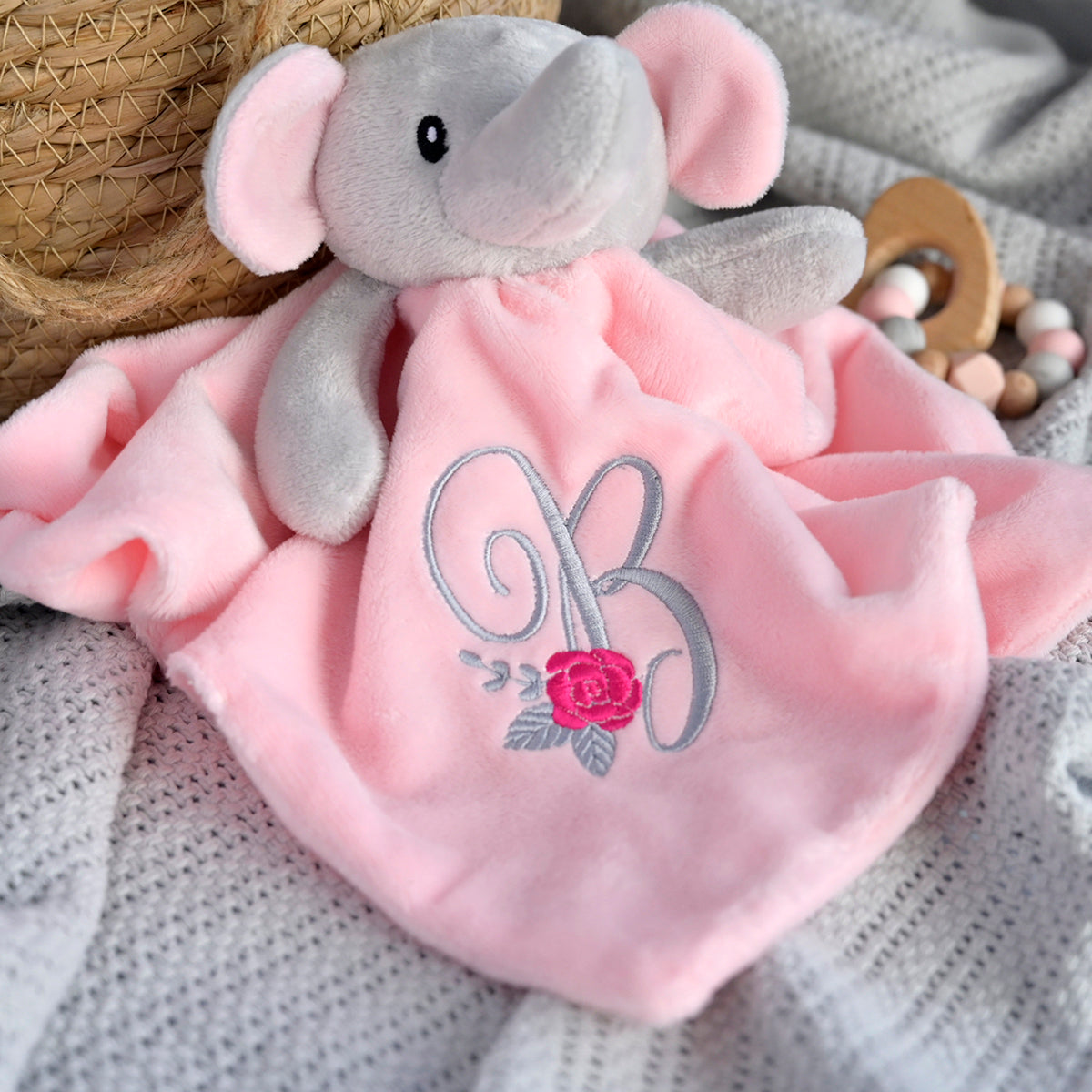 A plush elephant comforter with a soft pink blanket and embroidered monogram and rose, styled on a knitted blanket with a wooden rattle and wicker basket in the background.