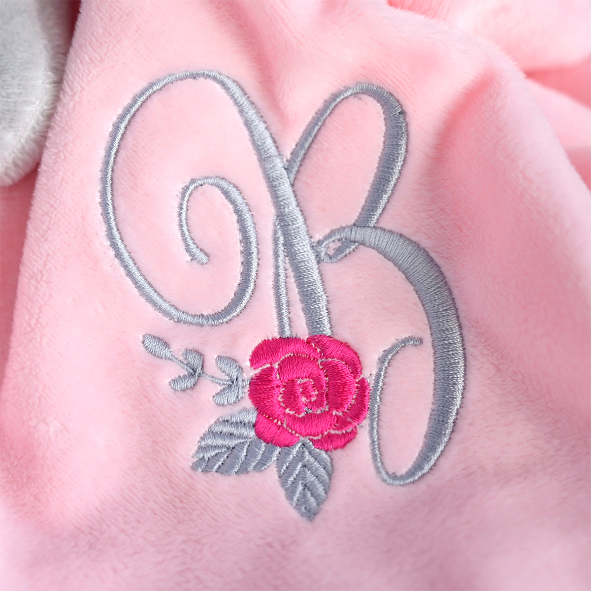 A close-up of a pink plush fabric featuring an embroidered monogram letter B in silver-gray with a bright pink rose and gray leaves.