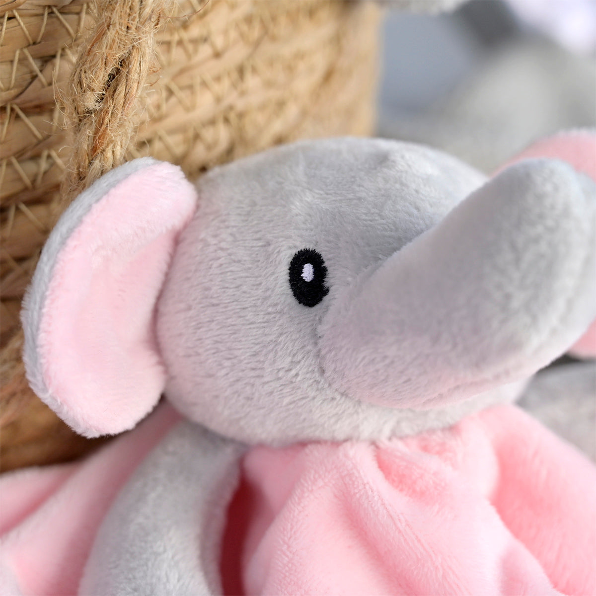 A close-up of a soft plush elephant comforter or stuffed toy with a pink blanket, featuring a gray face and trunk, pink ears, and a black embroidered eye.