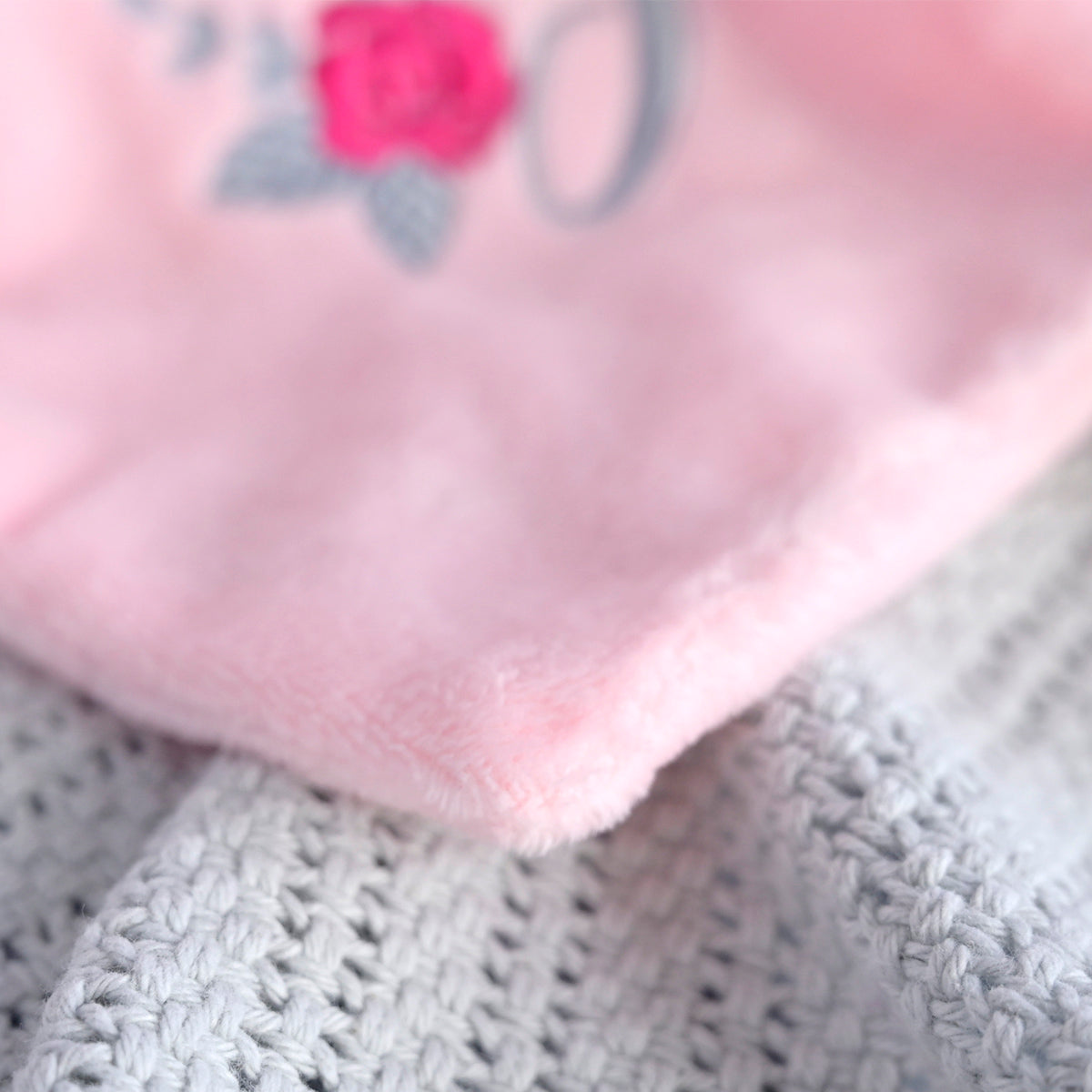A close-up of a soft pink plush fabric with a small embroidered pink rose and gray letter detail, resting on a light gray knitted textile.