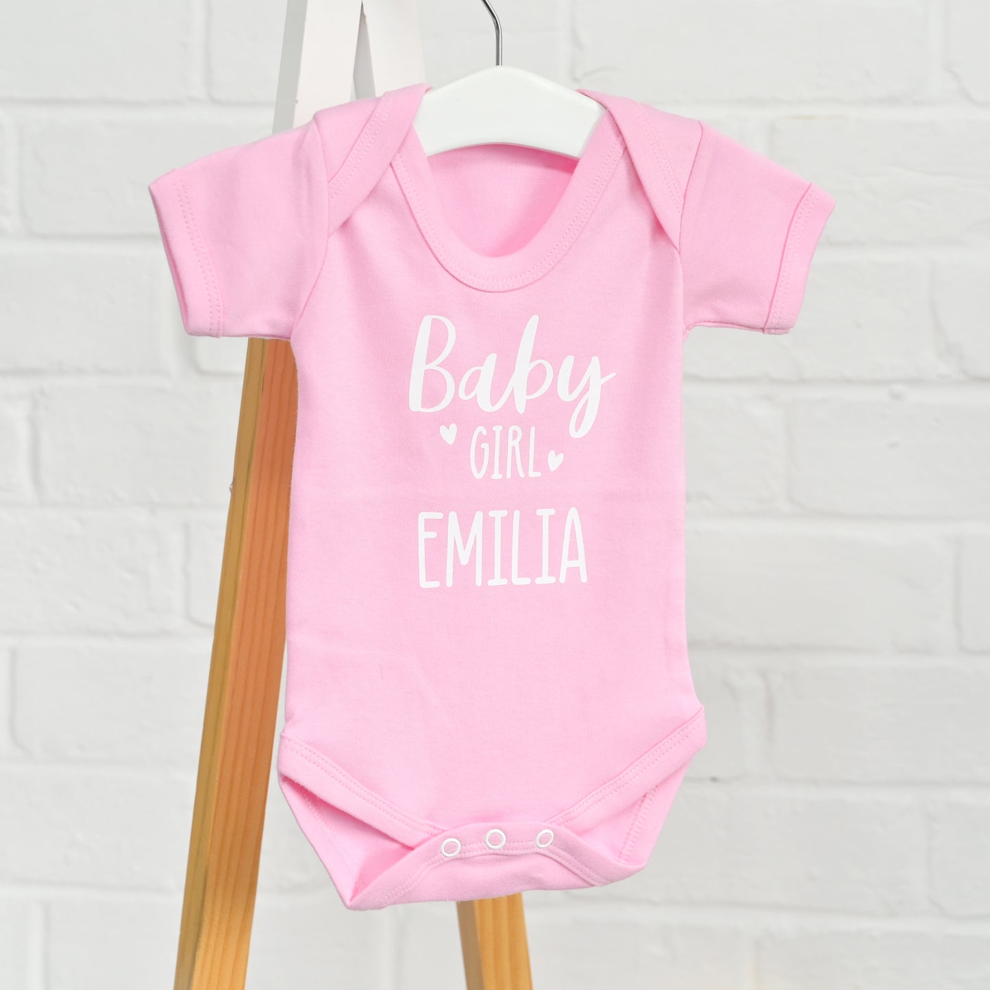 A light pink short-sleeve baby bodysuit hanging on a white hanger, with white text reading 'Baby Girl EMILIA' on the front, set against a white brick wall and wooden stand.