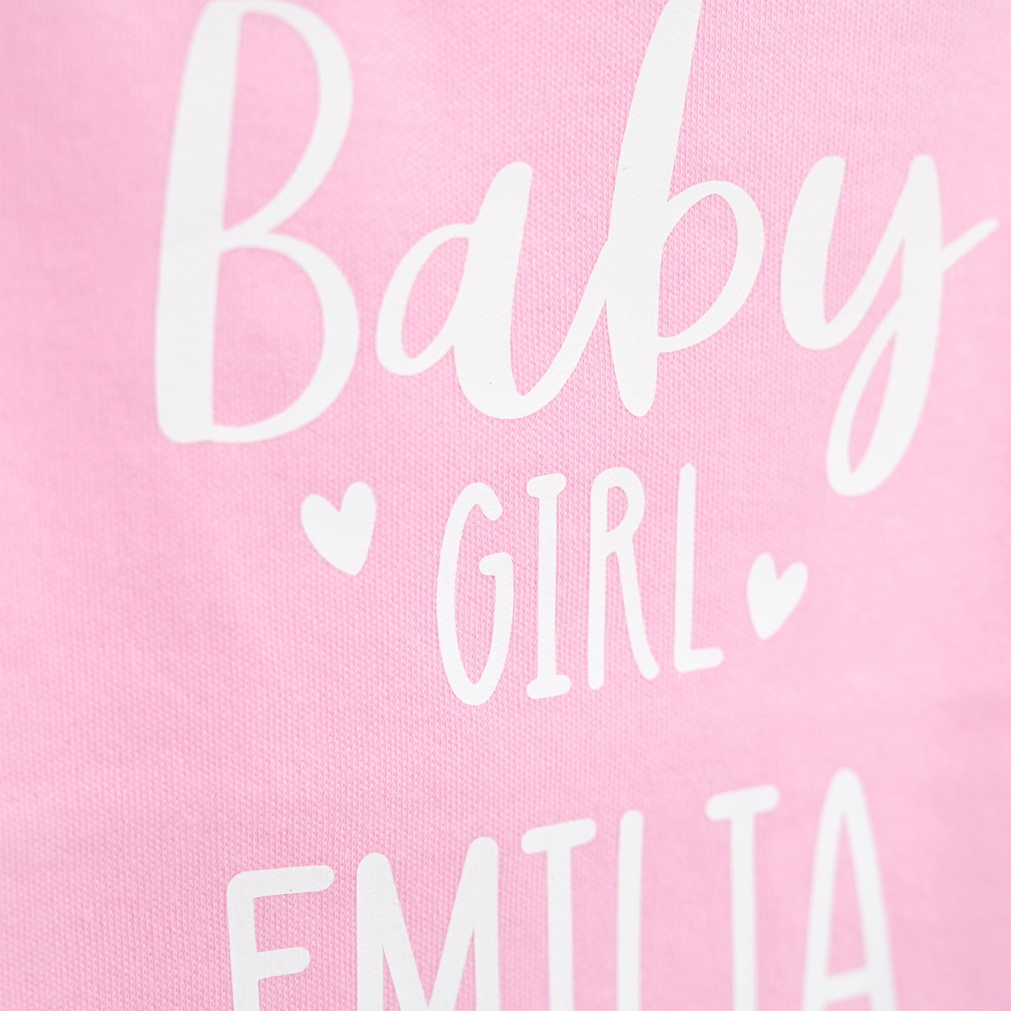 Close-up of a pink fabric surface with white printed text reading 'Baby GIRL' and part of 'EMILIA' below, plus small heart icons.
