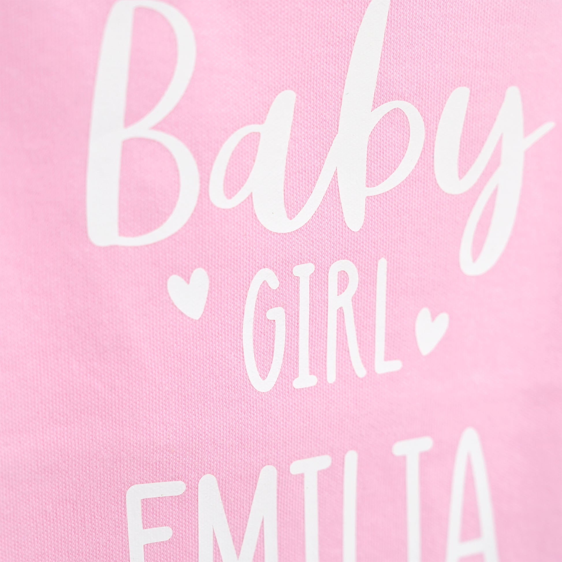 Close-up of a pink fabric surface with white printed text reading 'Baby GIRL' and part of 'EMILIA' below, plus small heart icons.