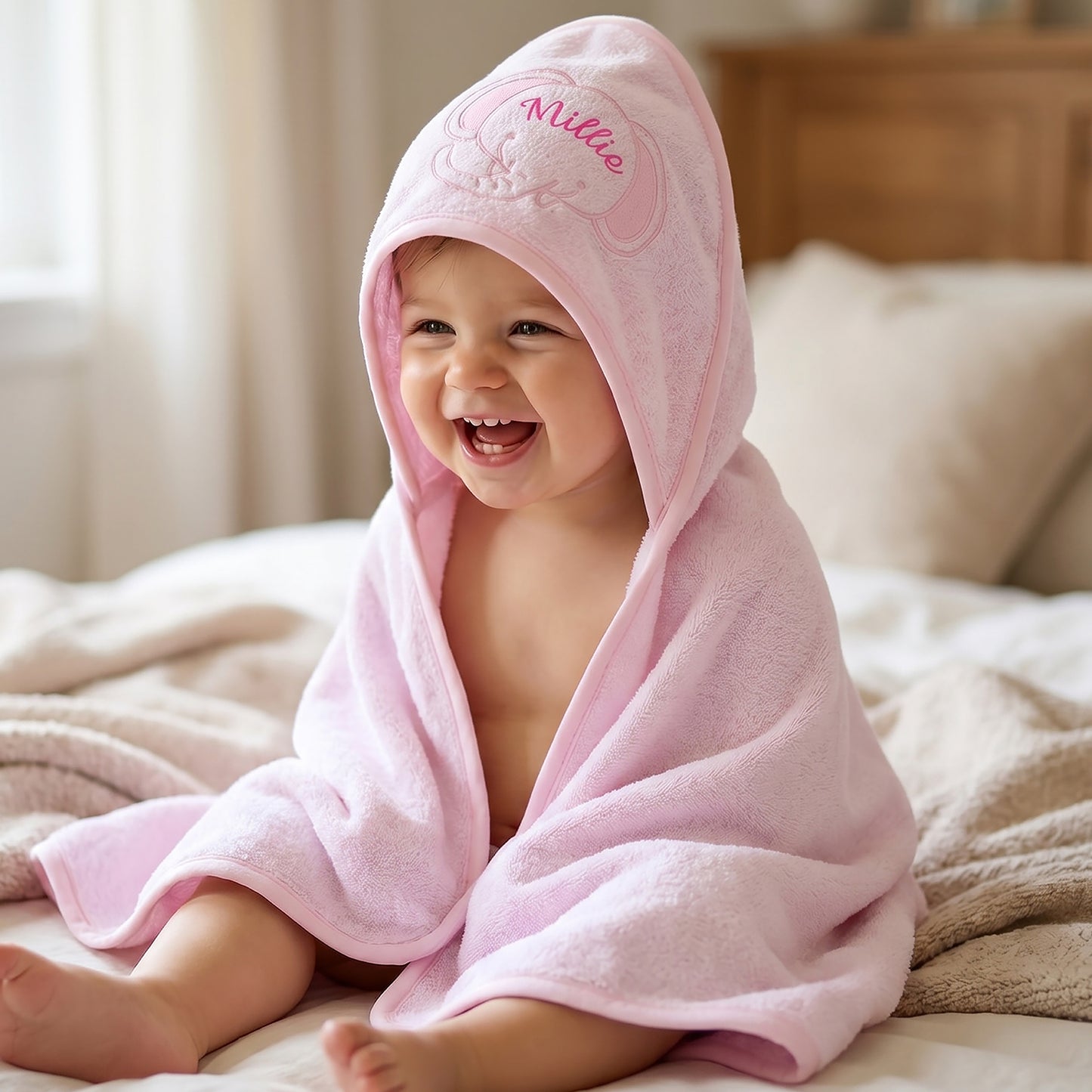 A smiling baby wrapped in a light pink hooded towel sits on a bed with neutral-toned bedding and a wooden headboard in the background. The hood is embroidered with the name 'Millie' in pink.