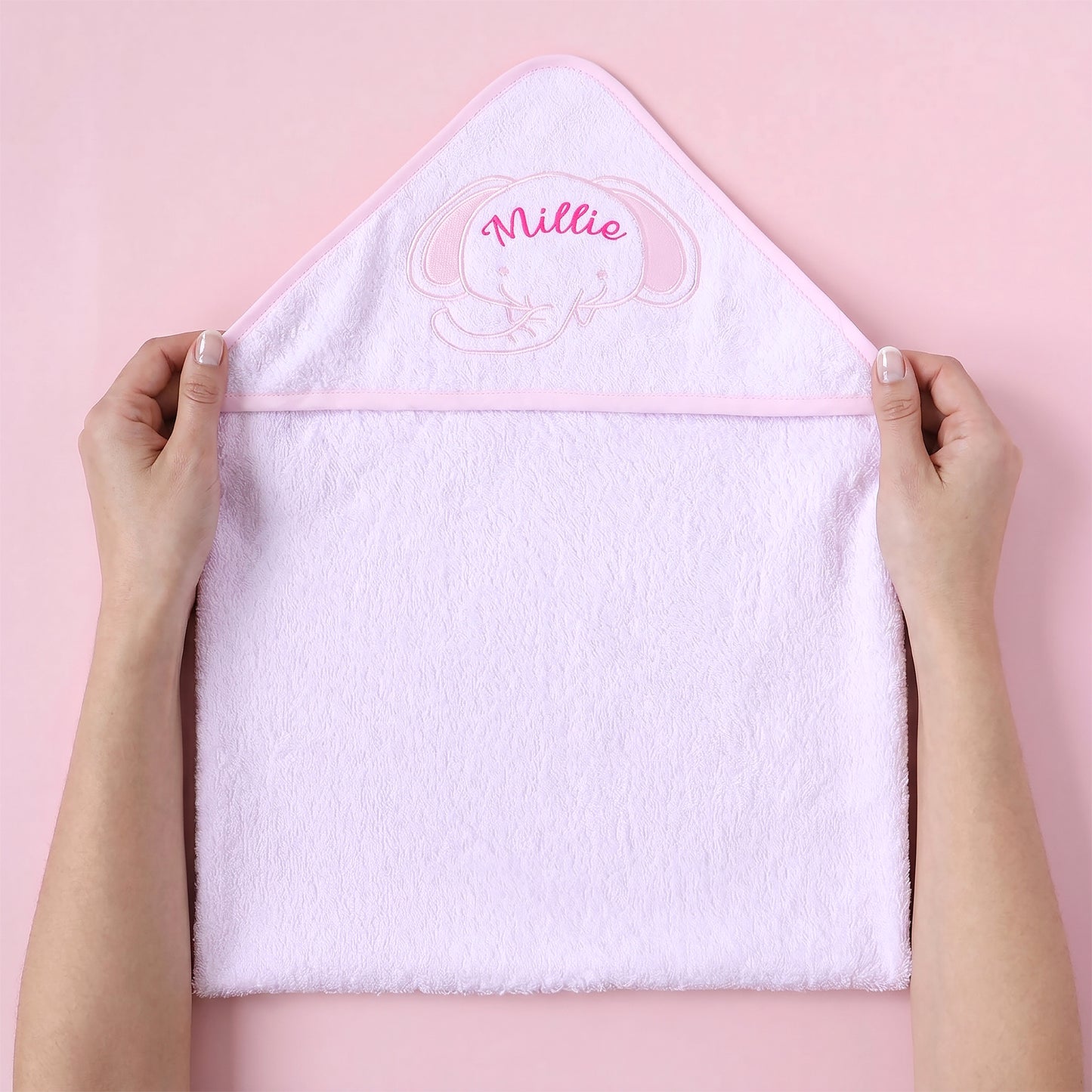 A light pink plush hooded towel held open by two hands against a solid pink background. The hood features an embroidered pink elephant face and the name 'Millie' in pink script.