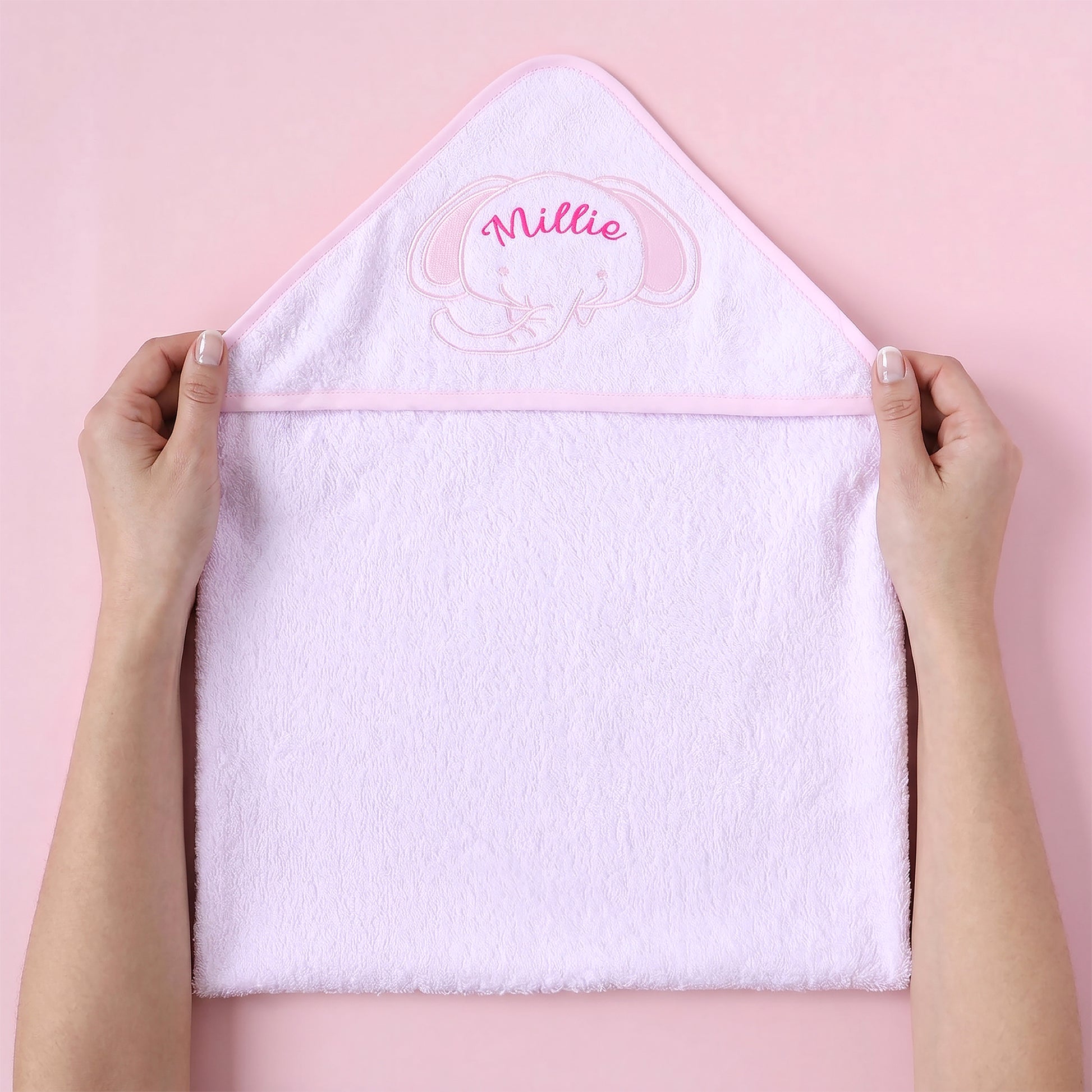 A light pink plush hooded towel held open by two hands against a solid pink background. The hood features an embroidered pink elephant face and the name 'Millie' in pink script.