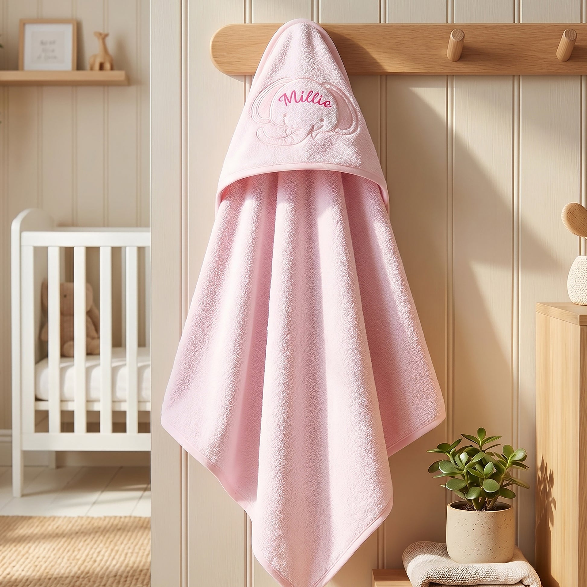 A soft pink baby hooded towel hangs from a wooden peg on a cream paneled wall in a styled nursery. The hood is embroidered with the name 'Millie' in pink script and a small bunny motif.