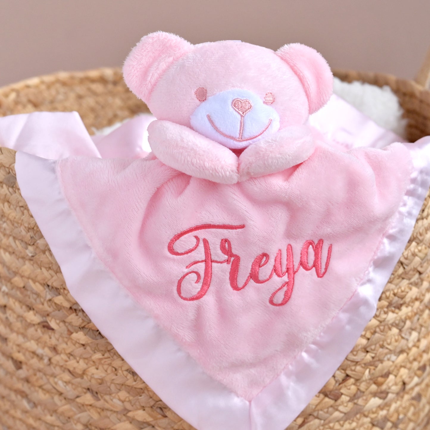 A pink plush teddy bear comforter blanket with a white satin border and the name 'Freya' embroidered in pink script, displayed inside a woven straw basket.