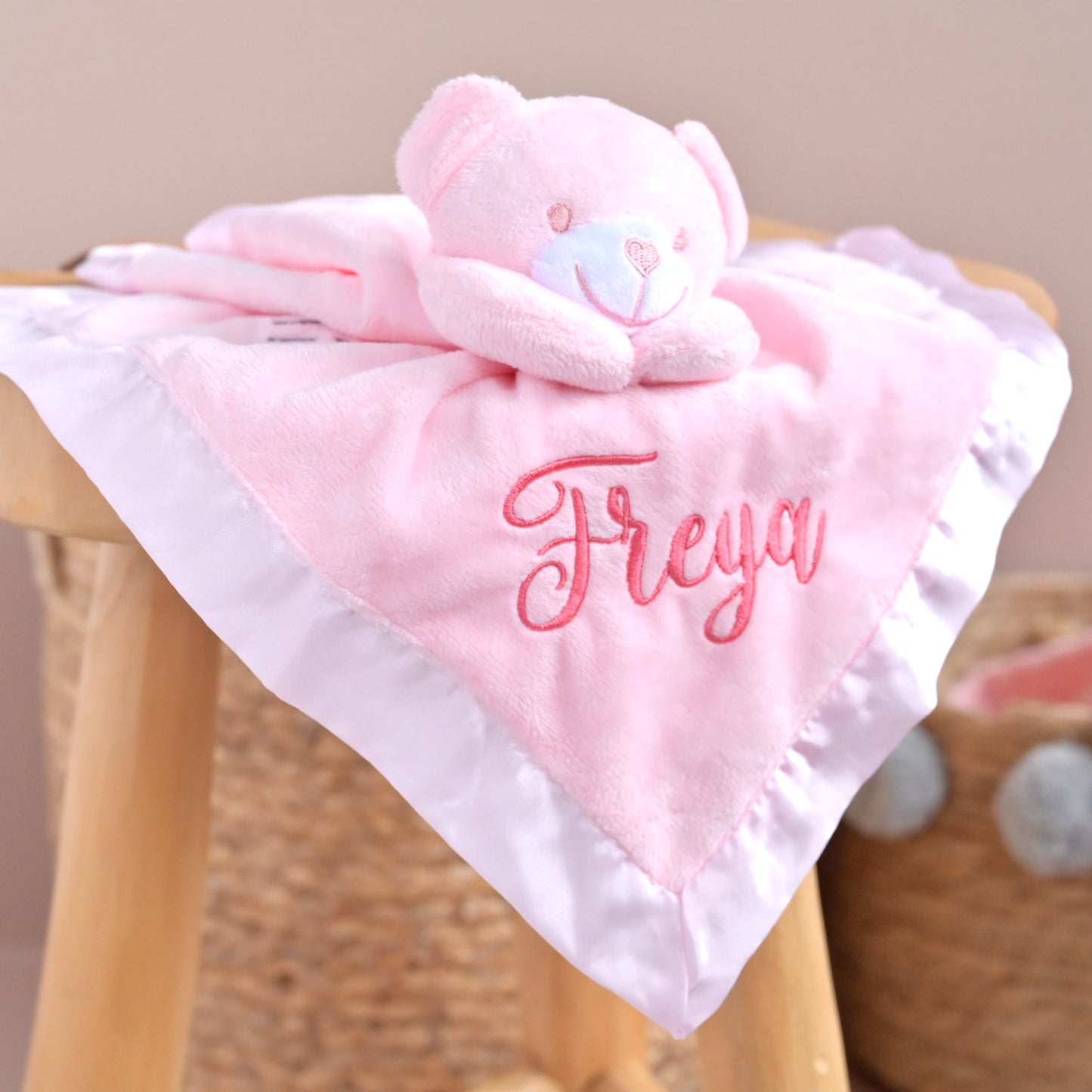 A pink plush bear comforter with a matching square blanket, draped over a wooden stool. The blanket features white ruffled trim and the name 'Freya' embroidered in pink script.